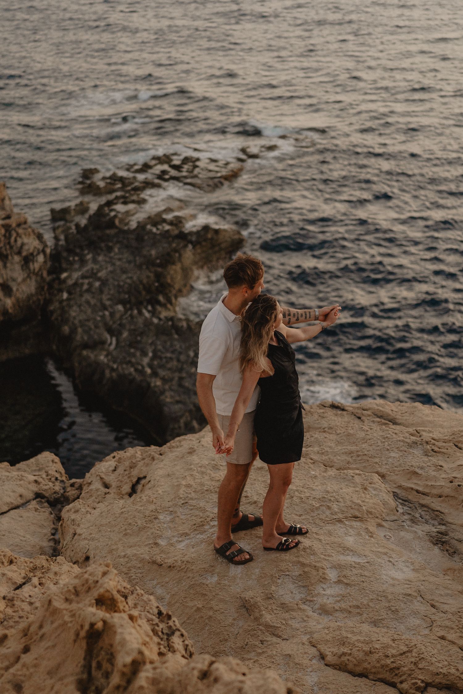 Couple standing on rocky coastal cliffs overlooking the ocean, sharing a romantic moment at sunset.
