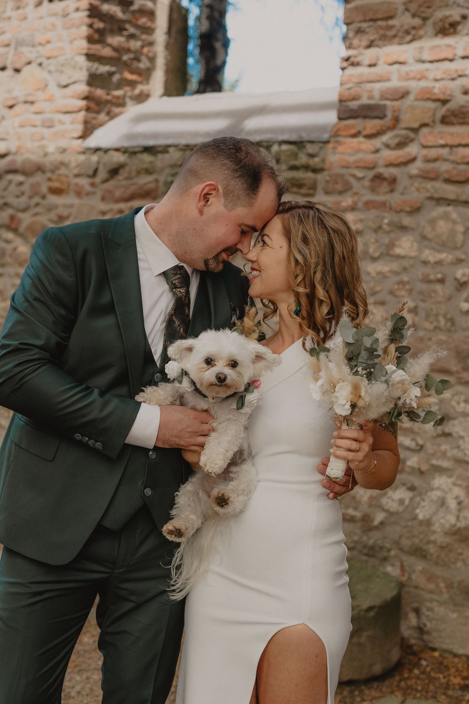A joyful wedding couple holding their small white dog and floral bouquet against a rustic stone wall.