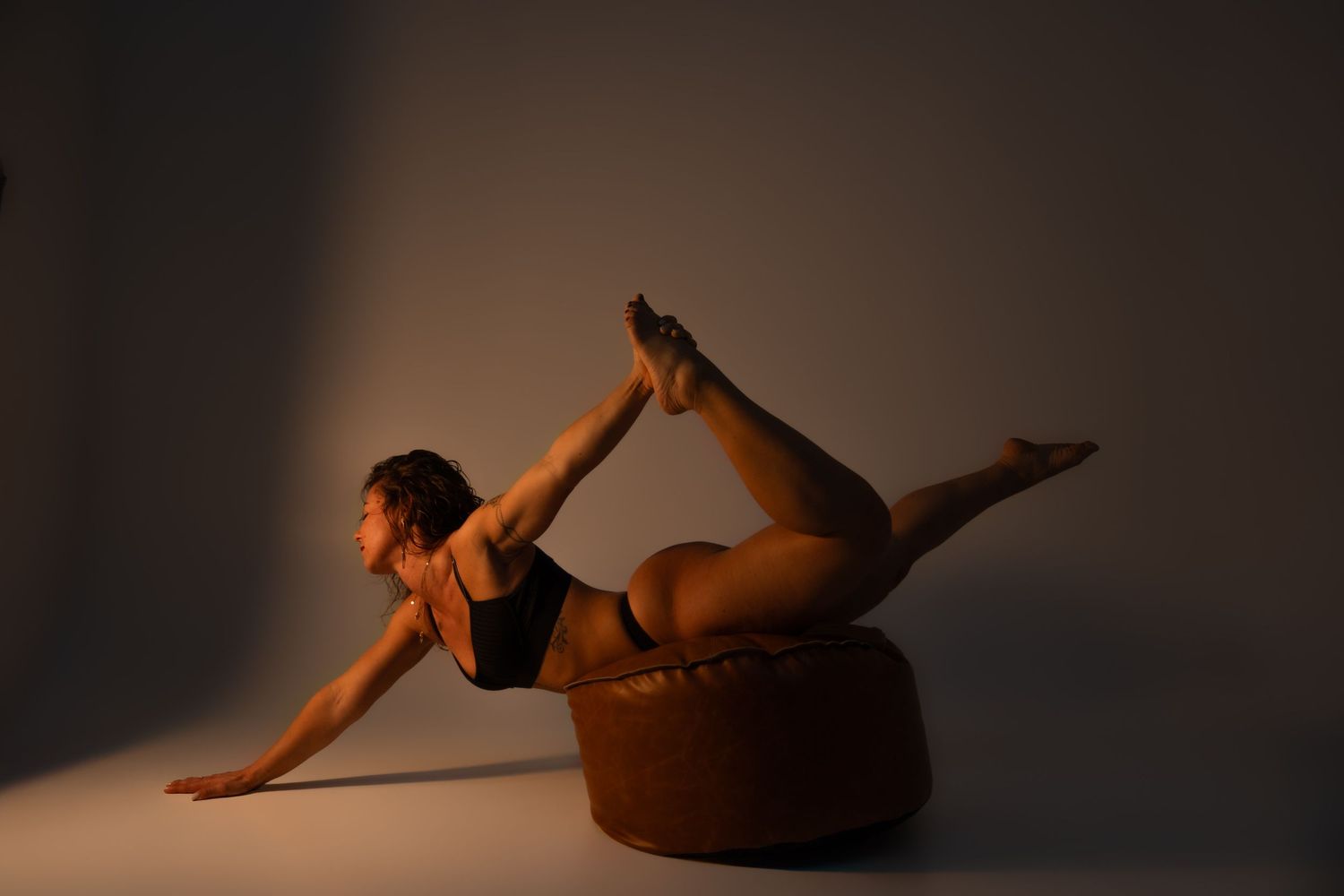 Flexible dancer balancing on a round ottoman in a black bodysuit, showcasing an impressive backbend pose in studio lighting.