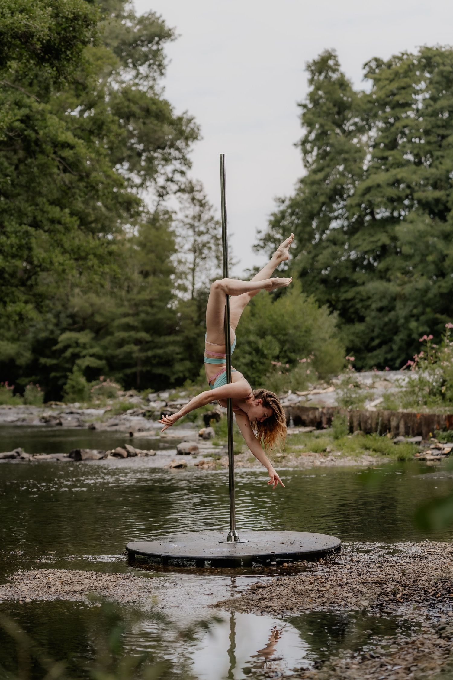 A woman performs an impressive pole dance move outdoors near a river, surrounded by lush green trees.
