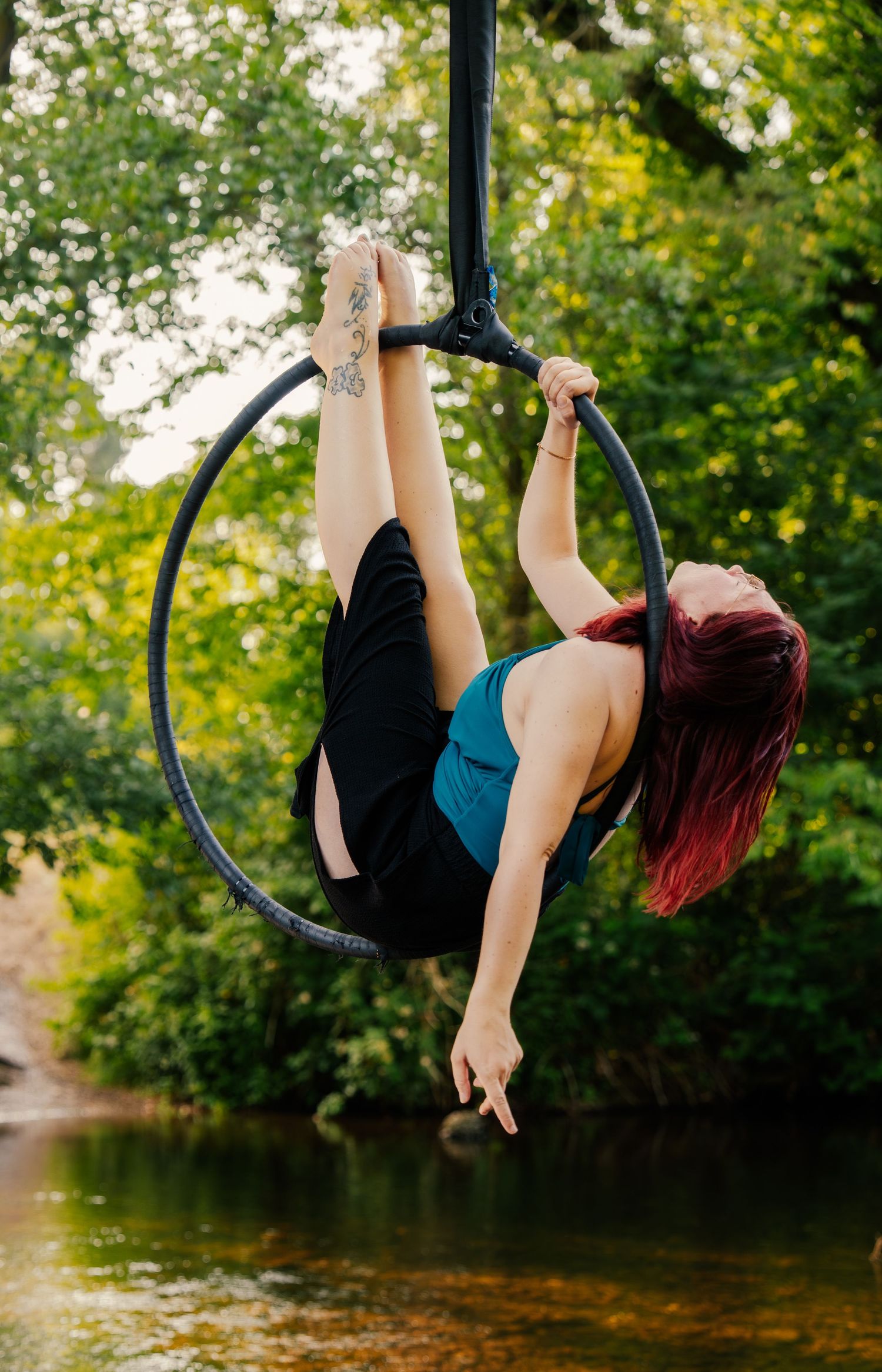 Woman with red hair performs aerial hoop lyra act outdoors over a stream surrounded by lush green trees.