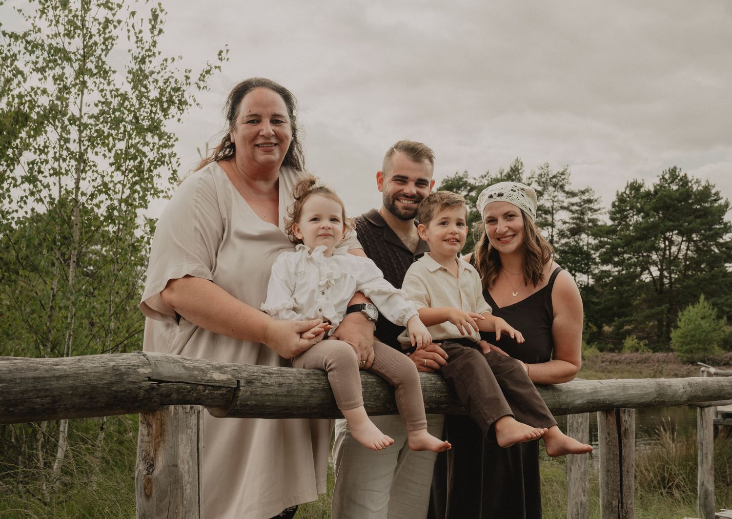 Family of five posing on a wooden fence outdoors, including two adults, two young children, and a smiling woman.