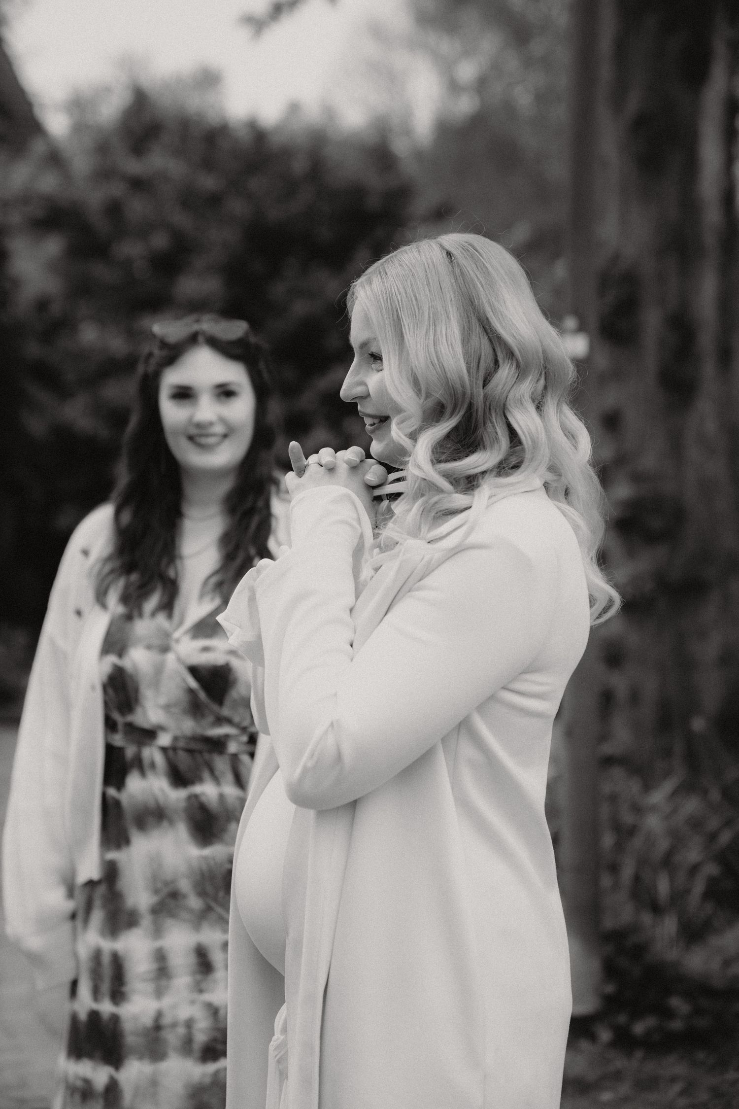 Black and white photo of a smiling pregnant woman in white coat with a friend in a garden.