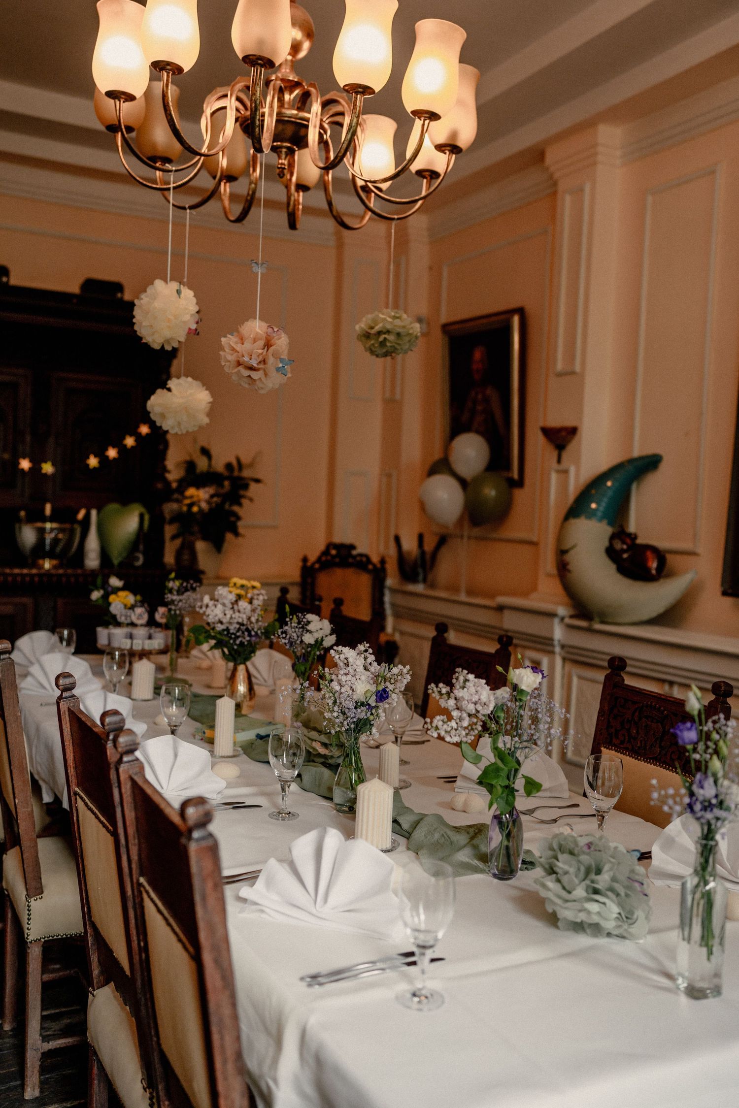 Elegant wedding reception table setting with white linens, floral centerpieces, candles, and a chandelier adorned with hanging pom-poms.