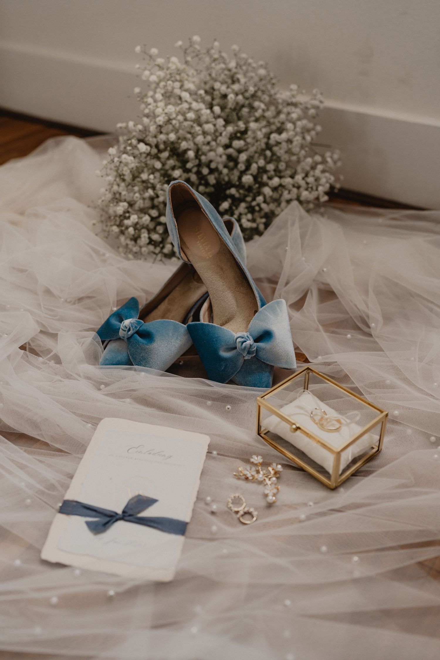 Wedding accessories including blue bow shoes, rings in a glass box, and baby's breath flowers on tulle fabric.