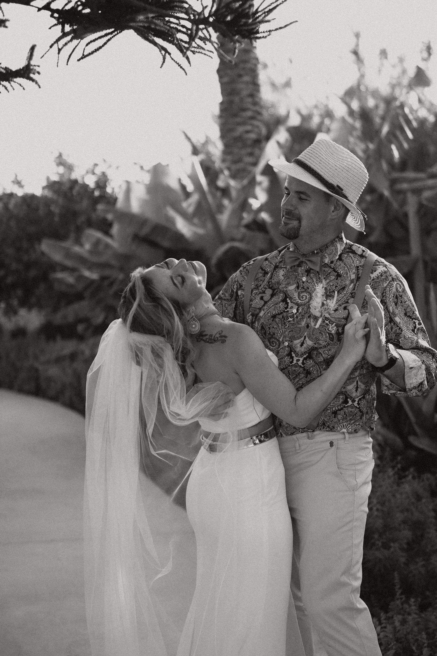A couple in wedding attire share a playful moment together outdoors with palm trees in the background.