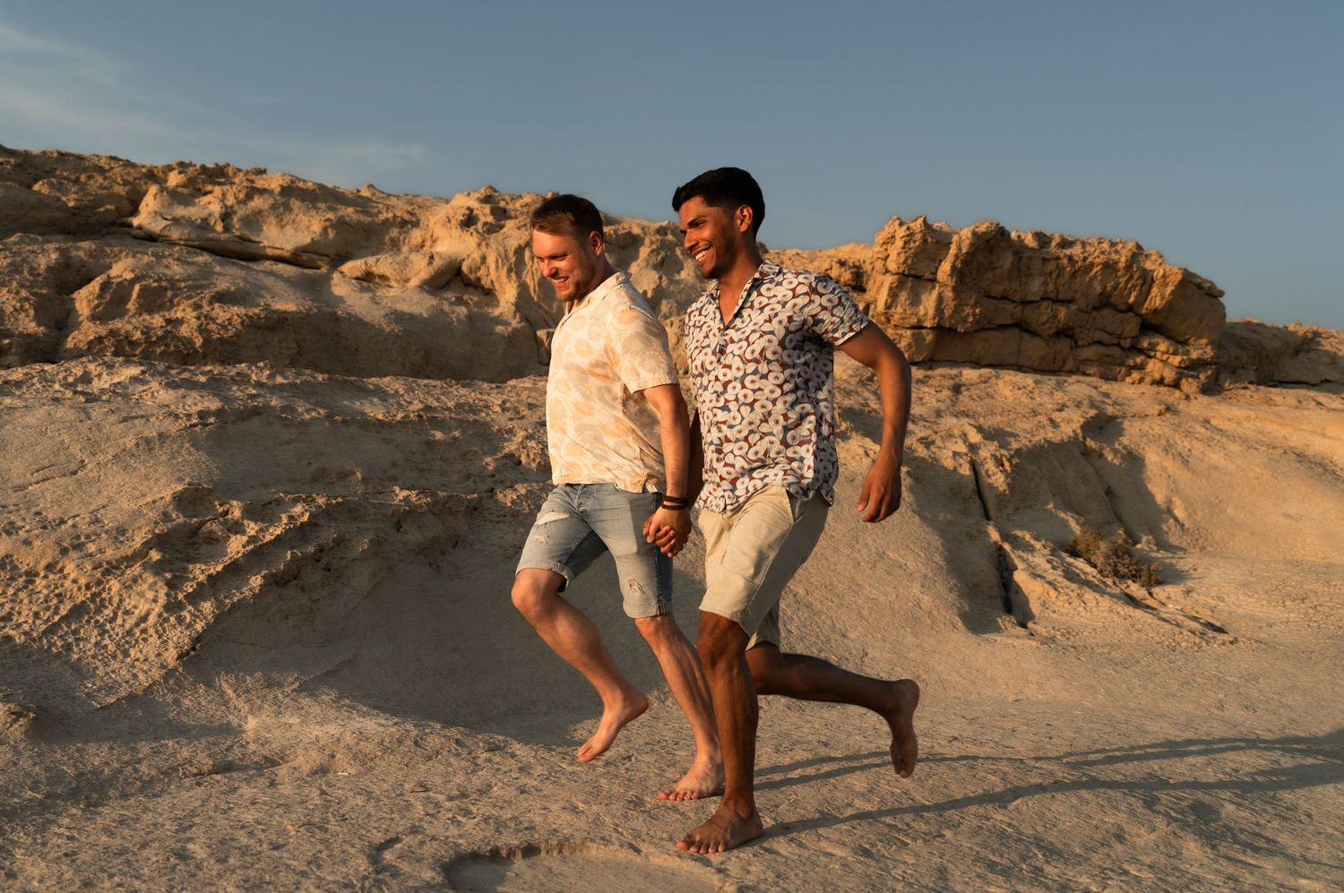 Two men walking barefoot on sandy beach rocks during golden hour sunset.