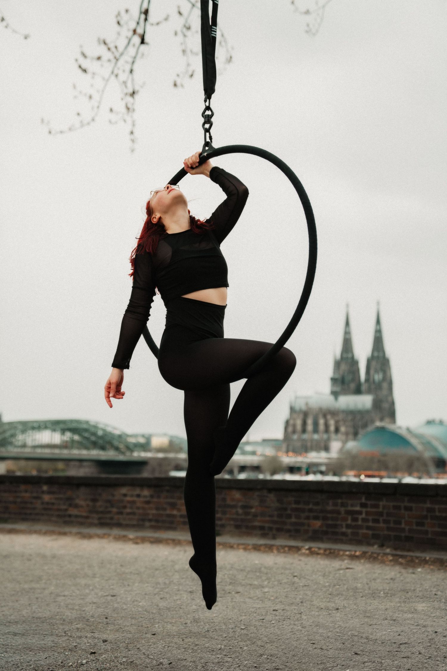 Red-haired aerial hoop dancer in black outfit performs outdoors with Cologne Cathedral in background.