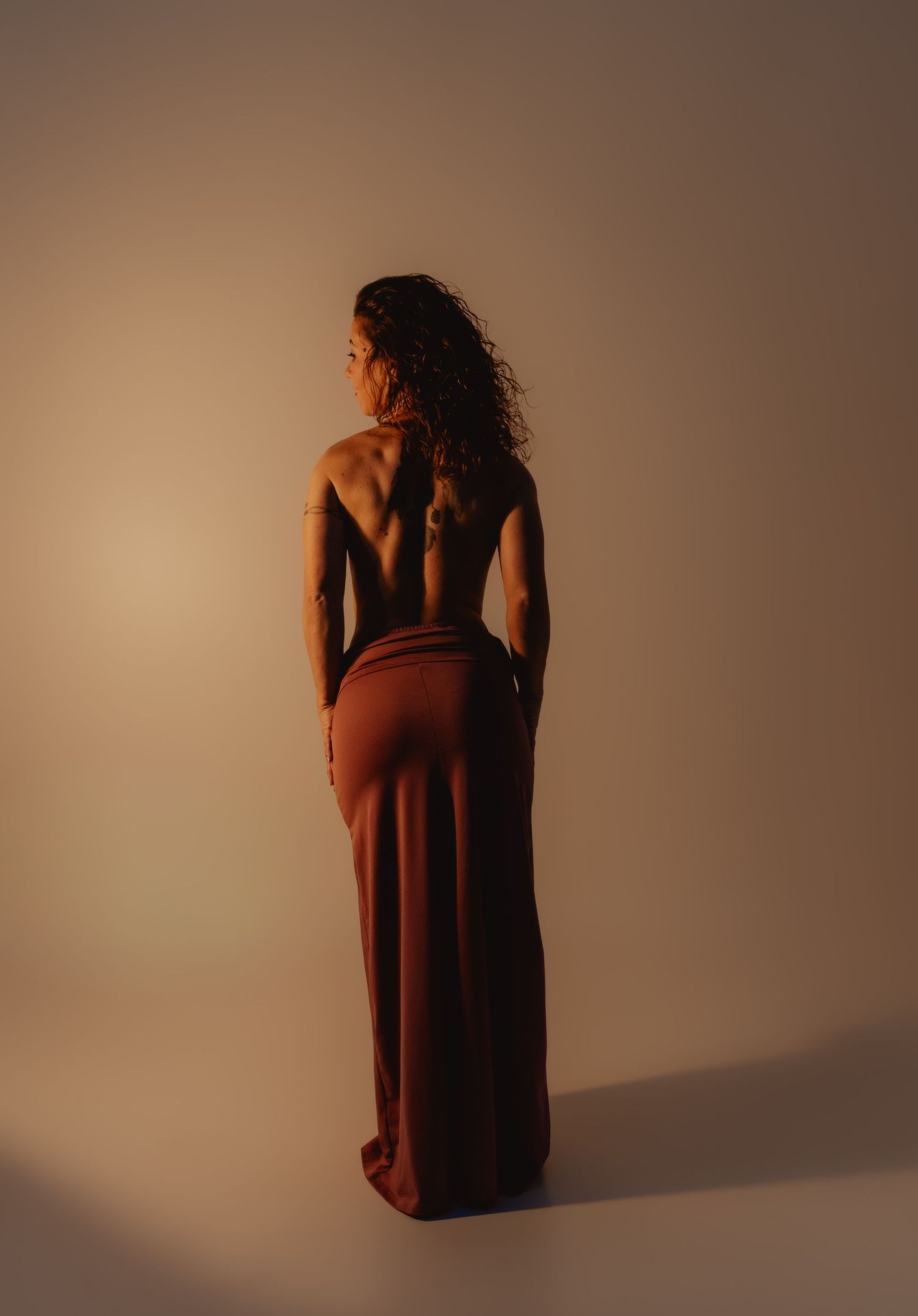 Woman in backless rust-colored halter gown poses gracefully, showcasing elegant open back design against neutral backdrop.