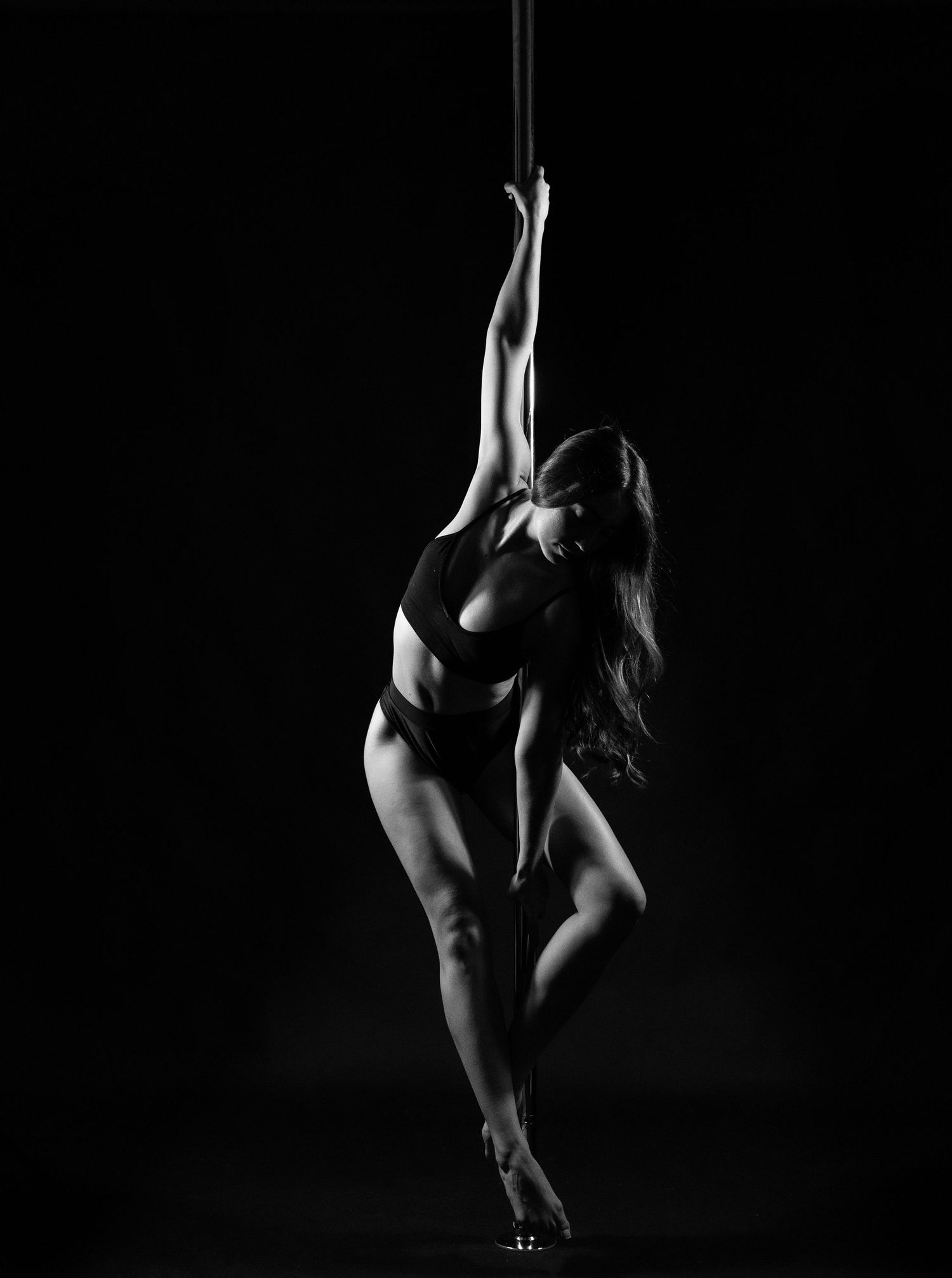 Black and white photo of a woman performing an elegant pole dance pose against a dark background.