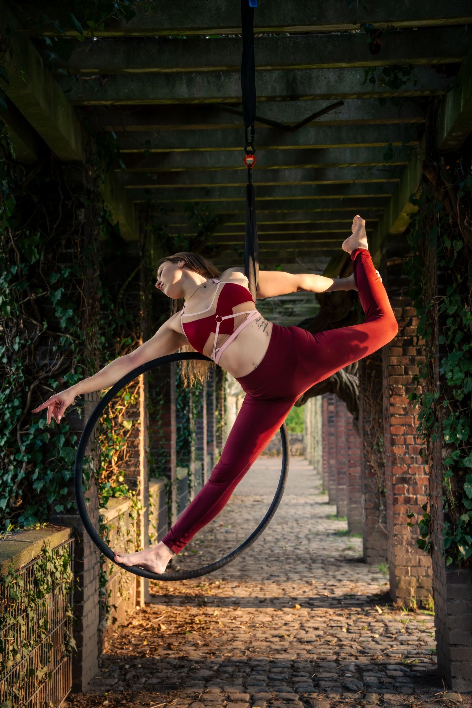 Aerial hoop artist in red performs a graceful split pose suspended in an ivy-covered brick archway on cobblestones.