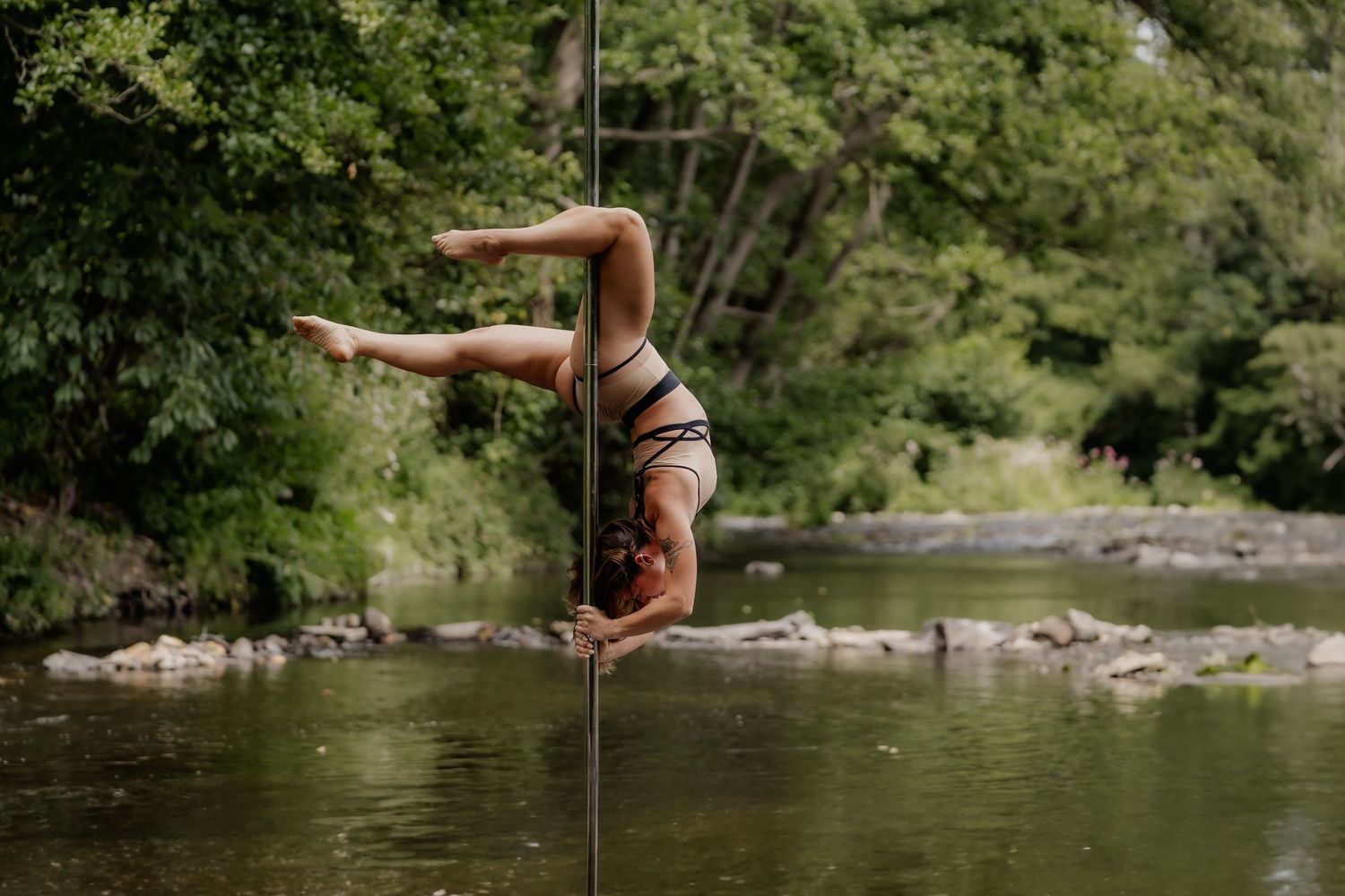 A woman performs an inverted pole dance move outdoors near a river surrounded by lush green trees.