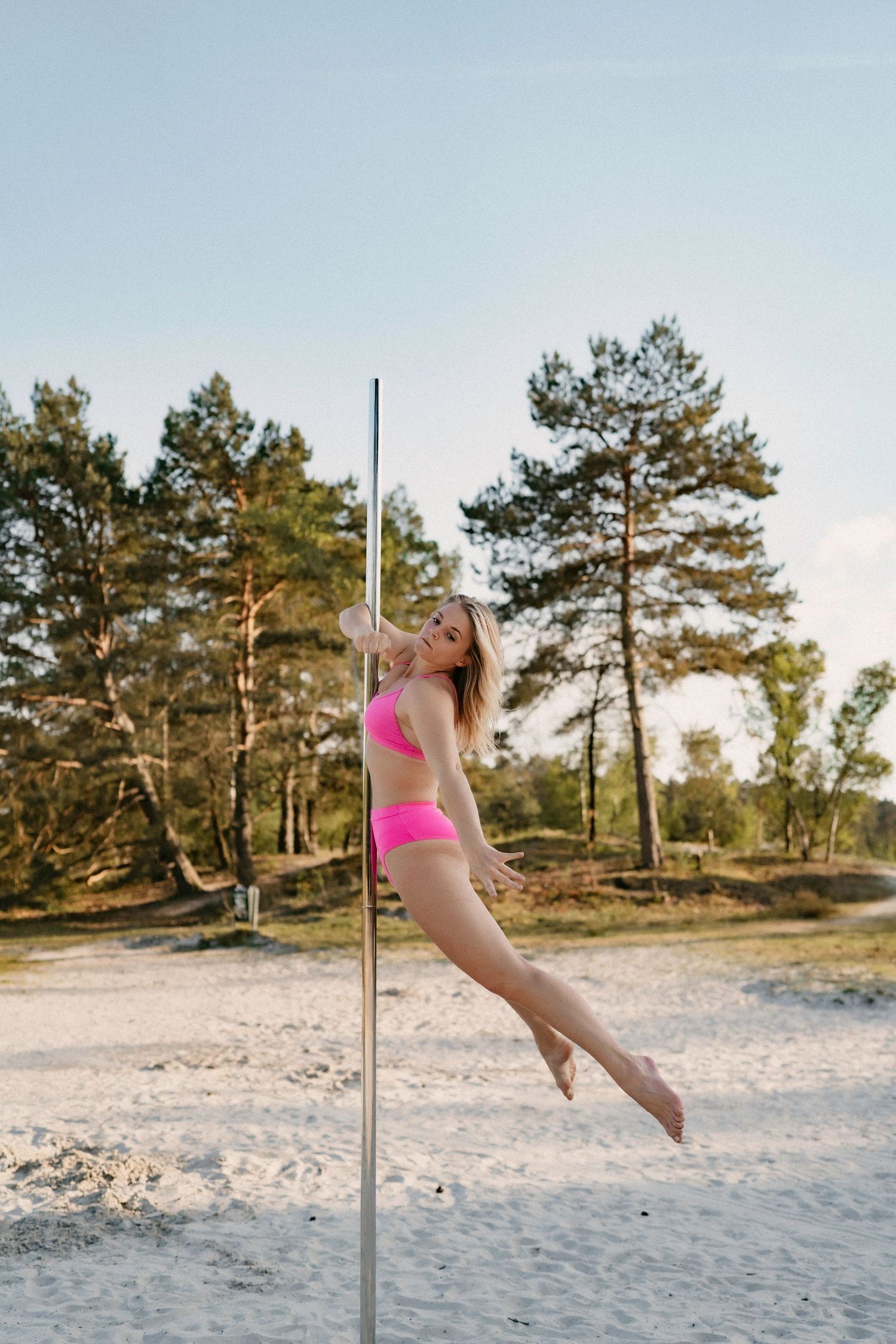 A blonde woman in a pink bikini performs pole dancing outdoors on a sandy area surrounded by pine trees.