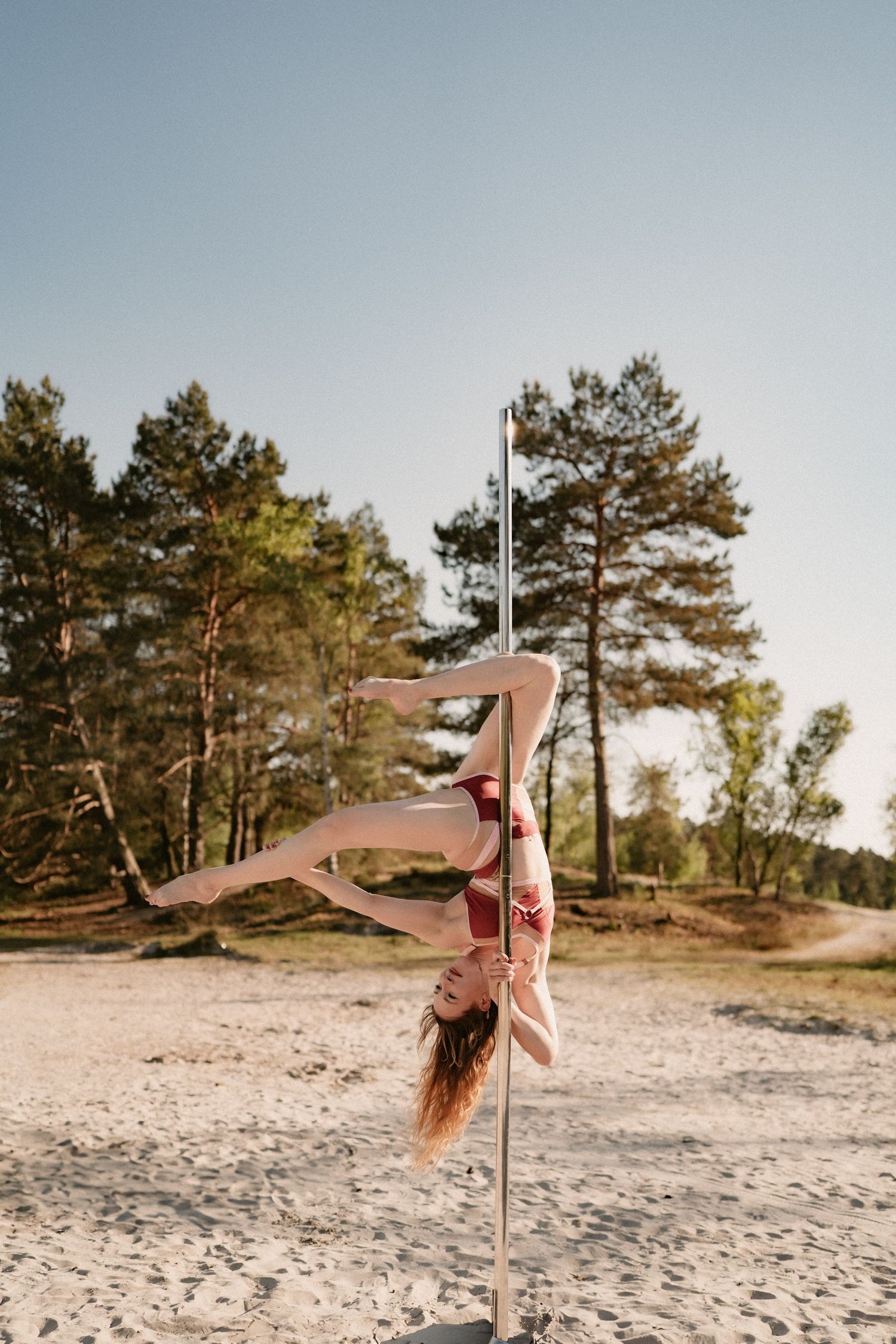 Woman performing an inverted pole dance move outdoors on sandy ground with pine trees in the background.