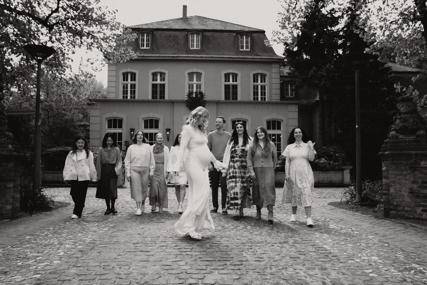Black and white photo of a bride in a white gown walking with her bridal party on cobblestones before an elegant estate.
