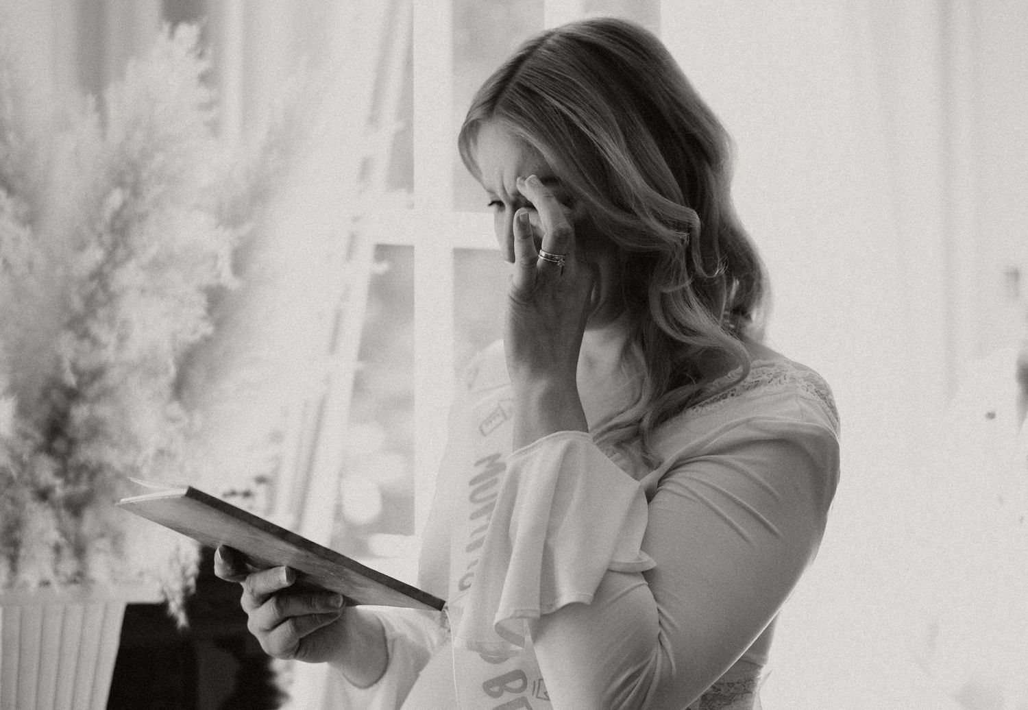 Black and white photo of an emotional woman in a ruffled dress reading a card by a window.