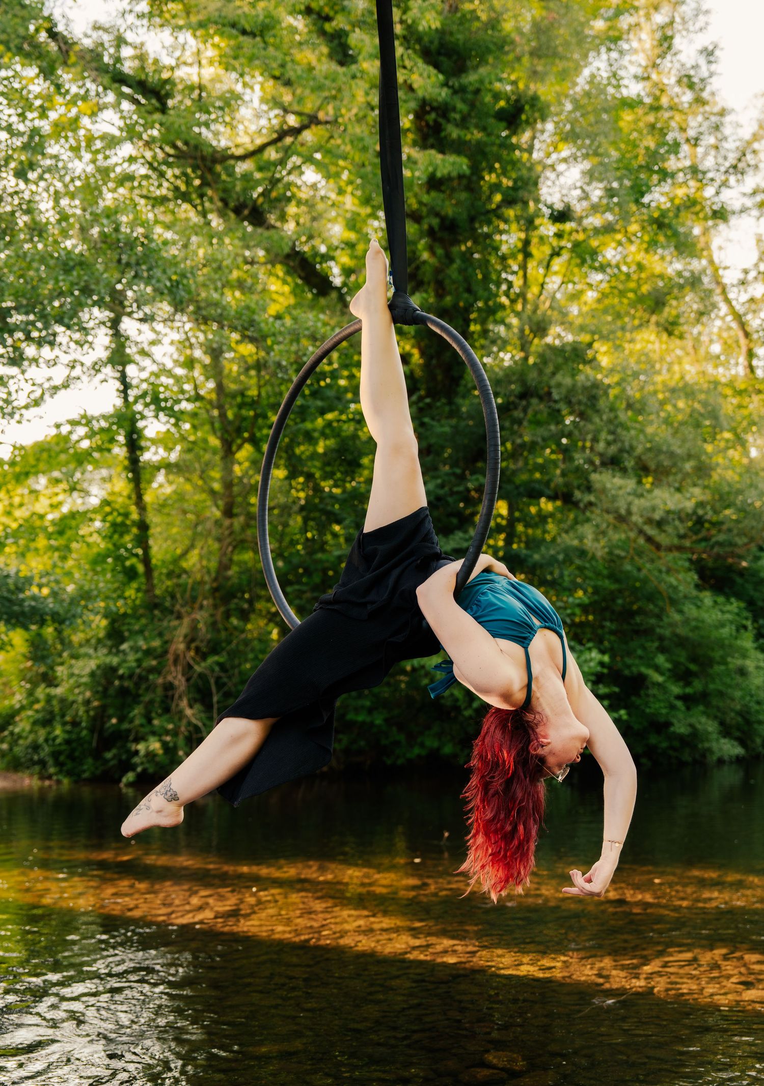 Red-haired aerial hoop performer executes a graceful backbend over a forest river, suspended from a lyra outdoors.
