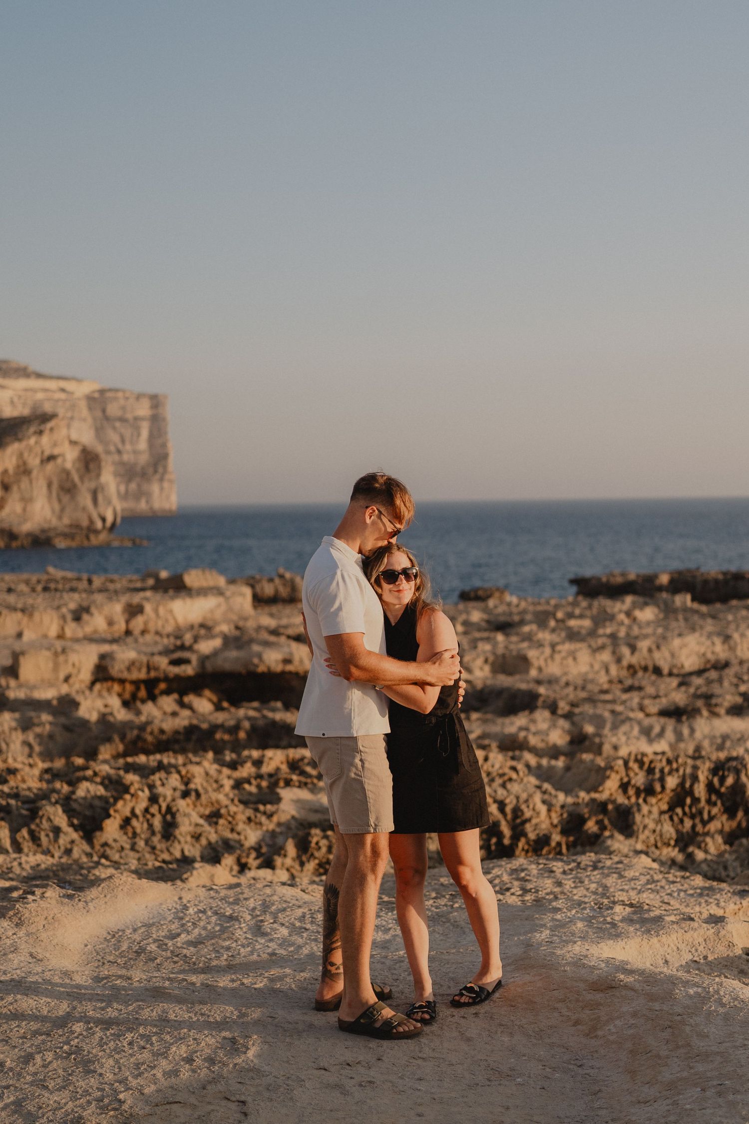 Couple embracing at sunset on rocky Mediterranean coastline with blue sea and cliffs in background.