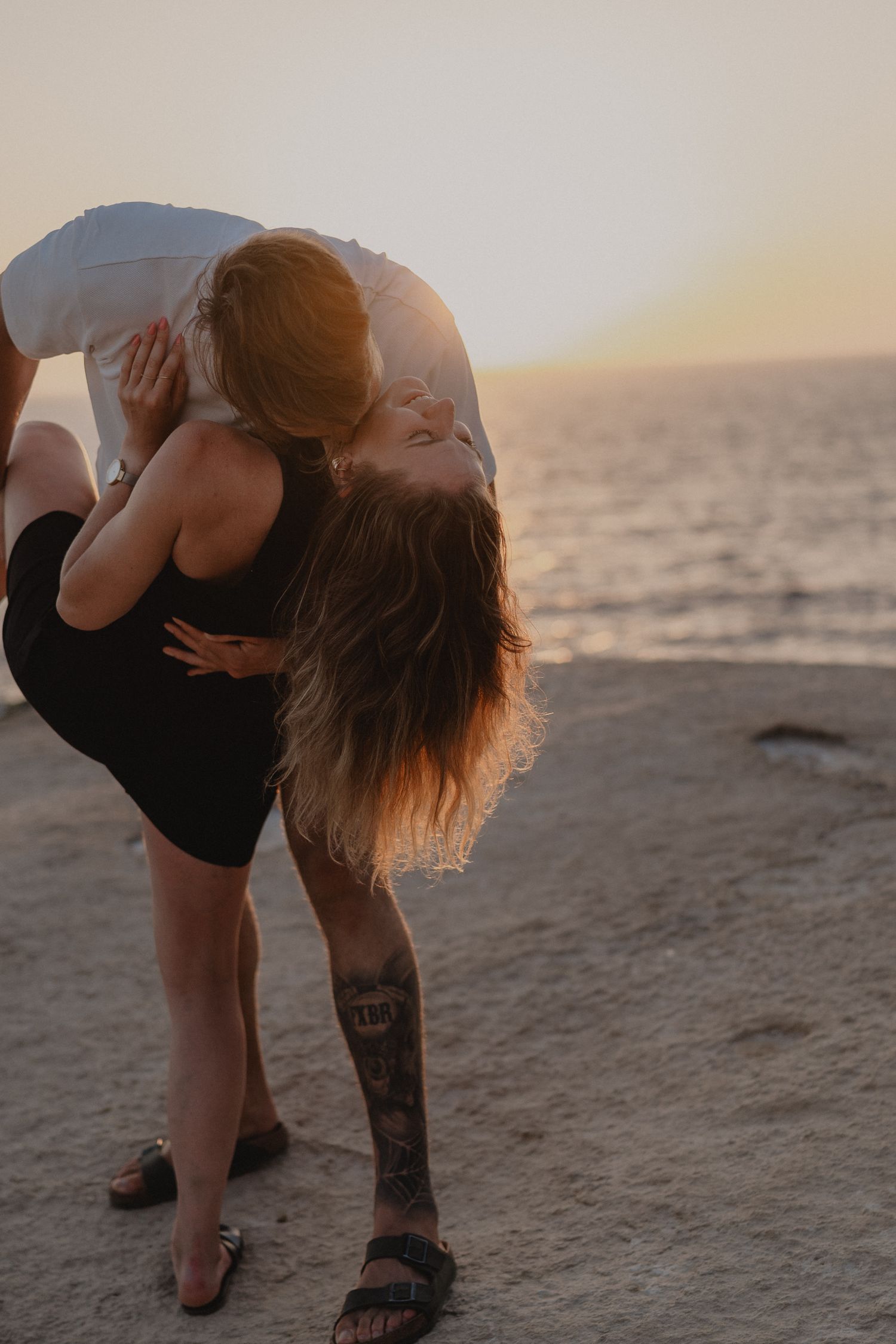 Couple sharing a romantic dip kiss on a rocky beach at sunset, woman with tattoo in black dress.