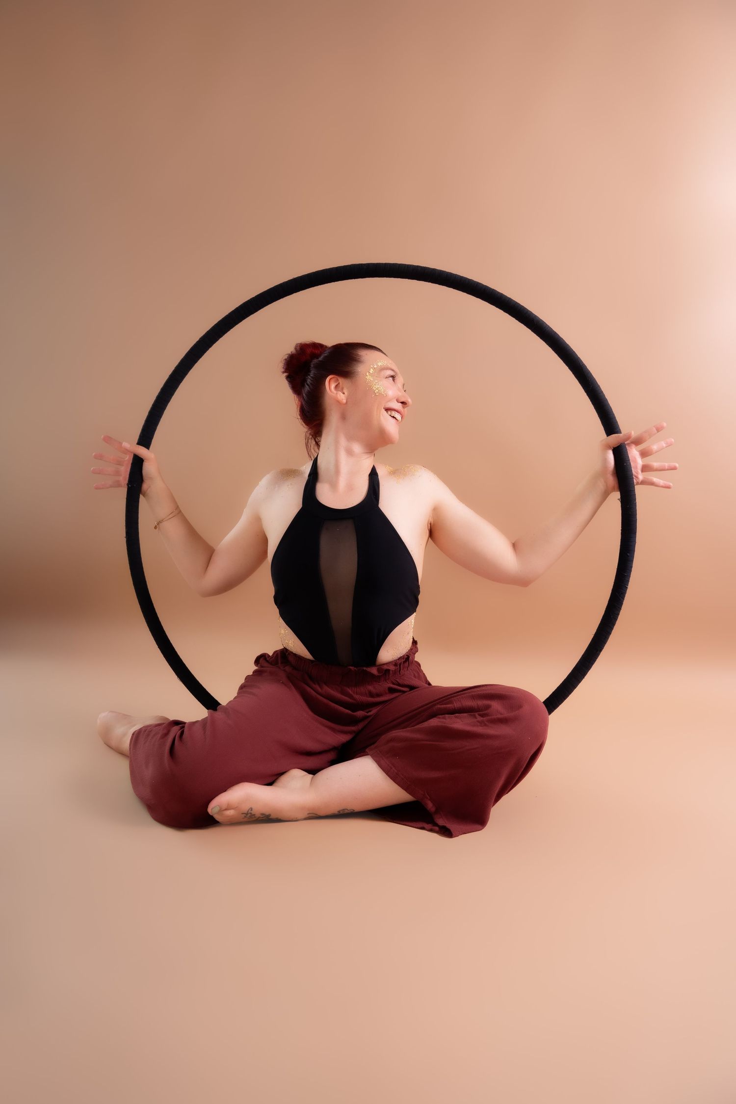 Smiling woman in black top and maroon pants sits cross-legged inside a large black hoop on a beige background.