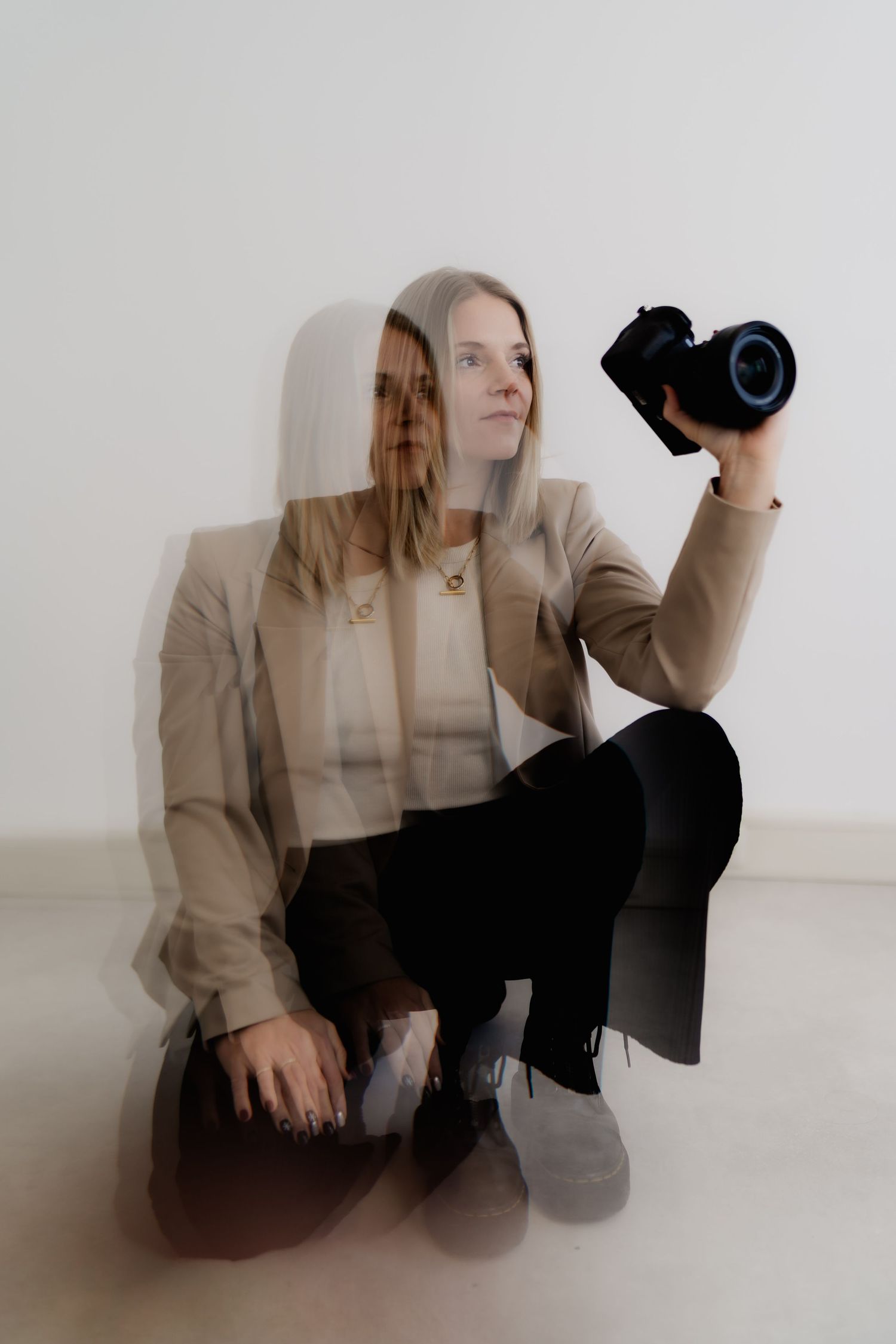 A person in a beige blazer and black pants poses with a camera in a minimalist white studio setting.