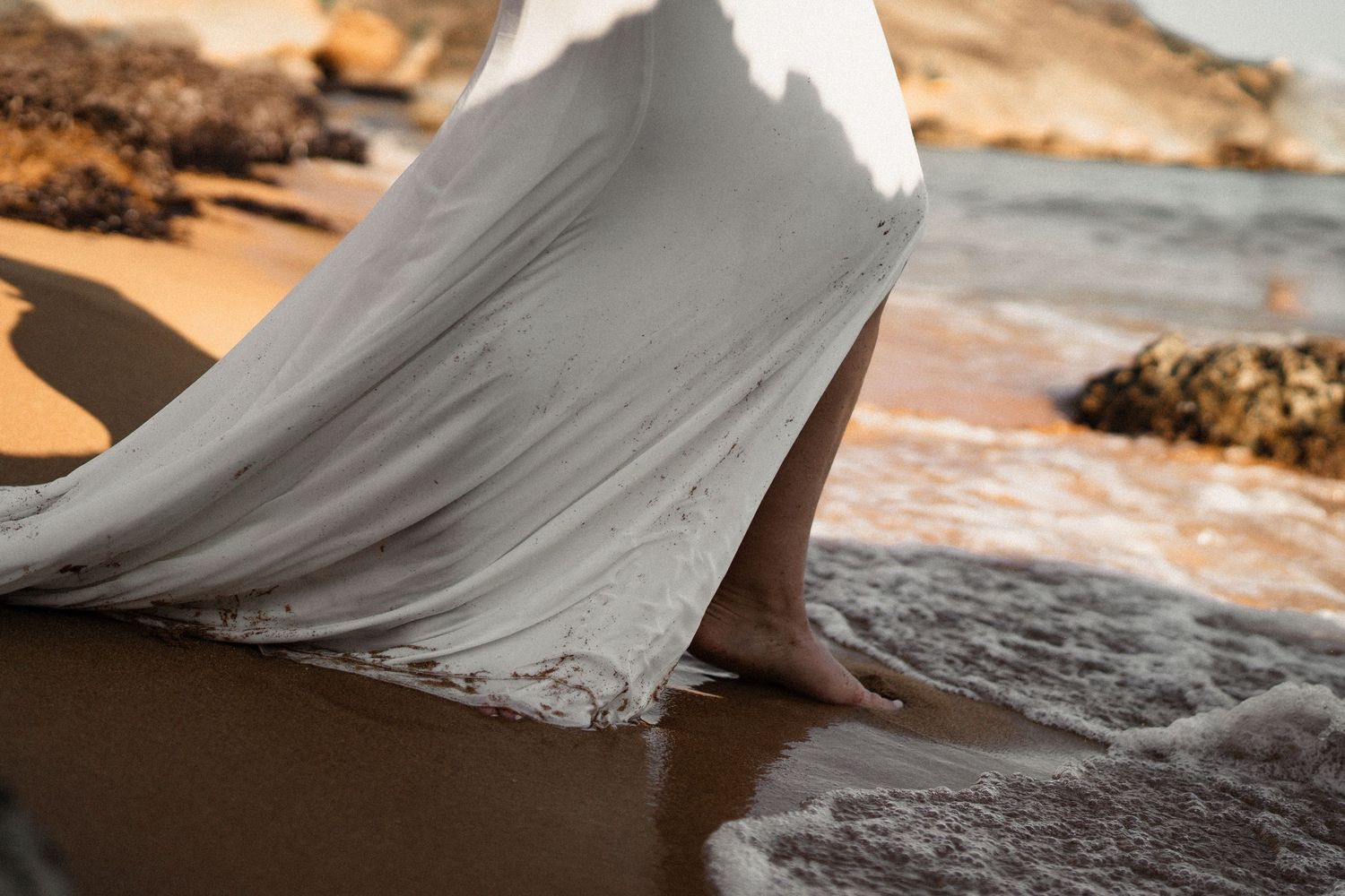Flowing white dress trails gracefully across wet sand during beach photoshoot.