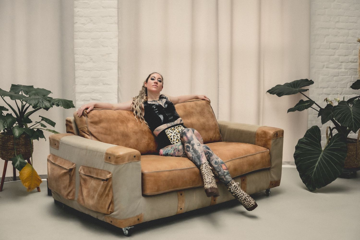 Woman with blonde braids reclines on a worn leather loveseat surrounded by tropical plants in a bright white studio.