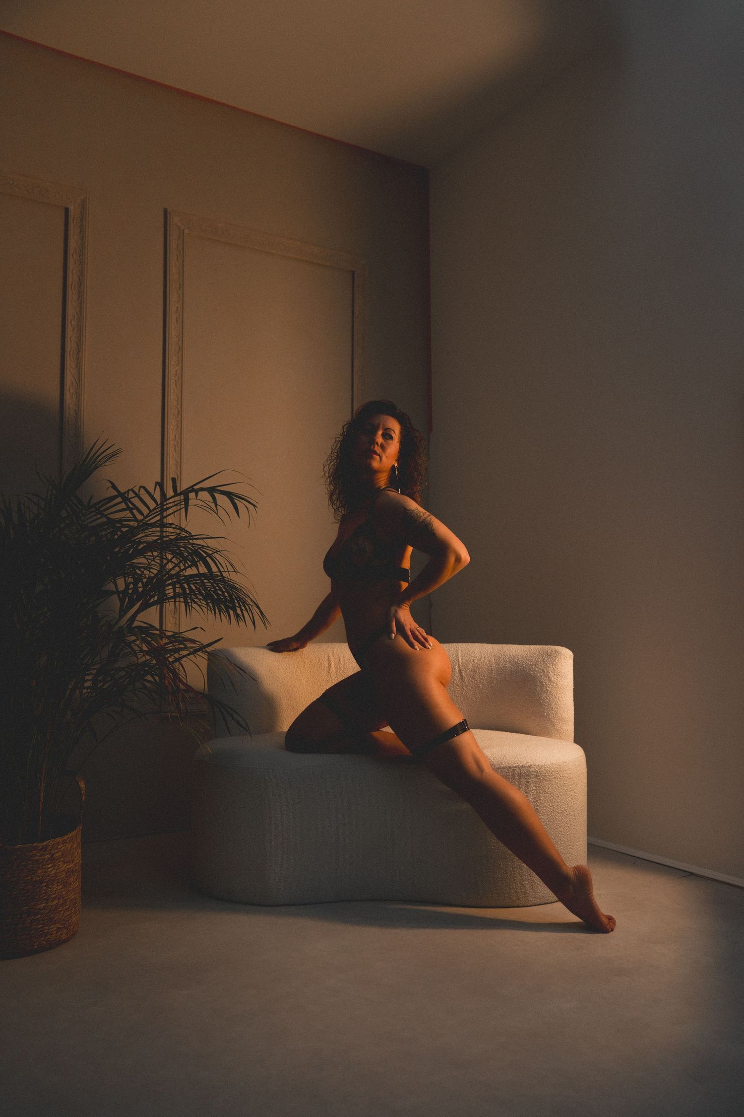 Woman in dark lingerie posing elegantly beside a white sofa with warm ambient lighting and a potted plant.