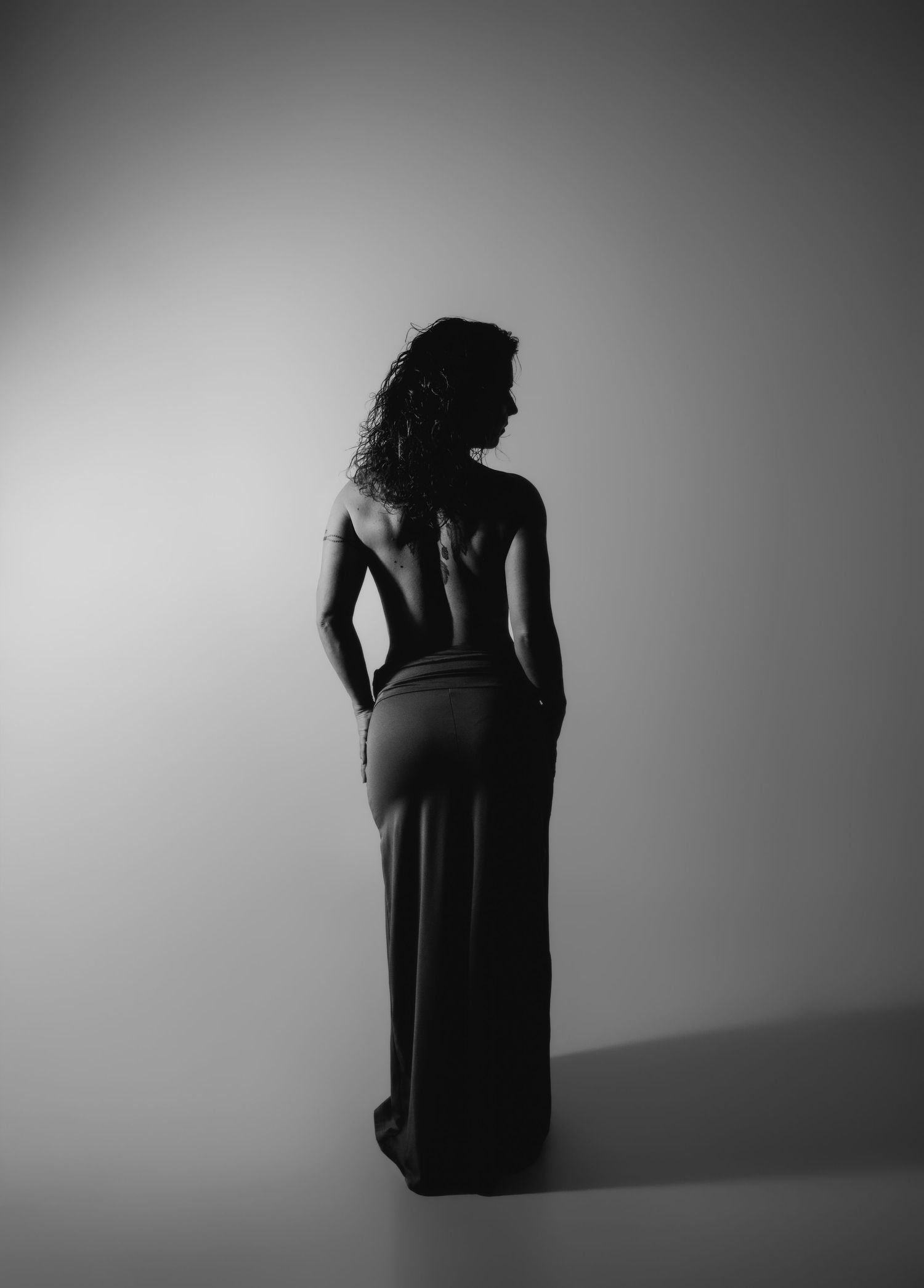 Black and white studio photo of a woman in a backless maxi skirt, showcasing her bare back and curly hair.