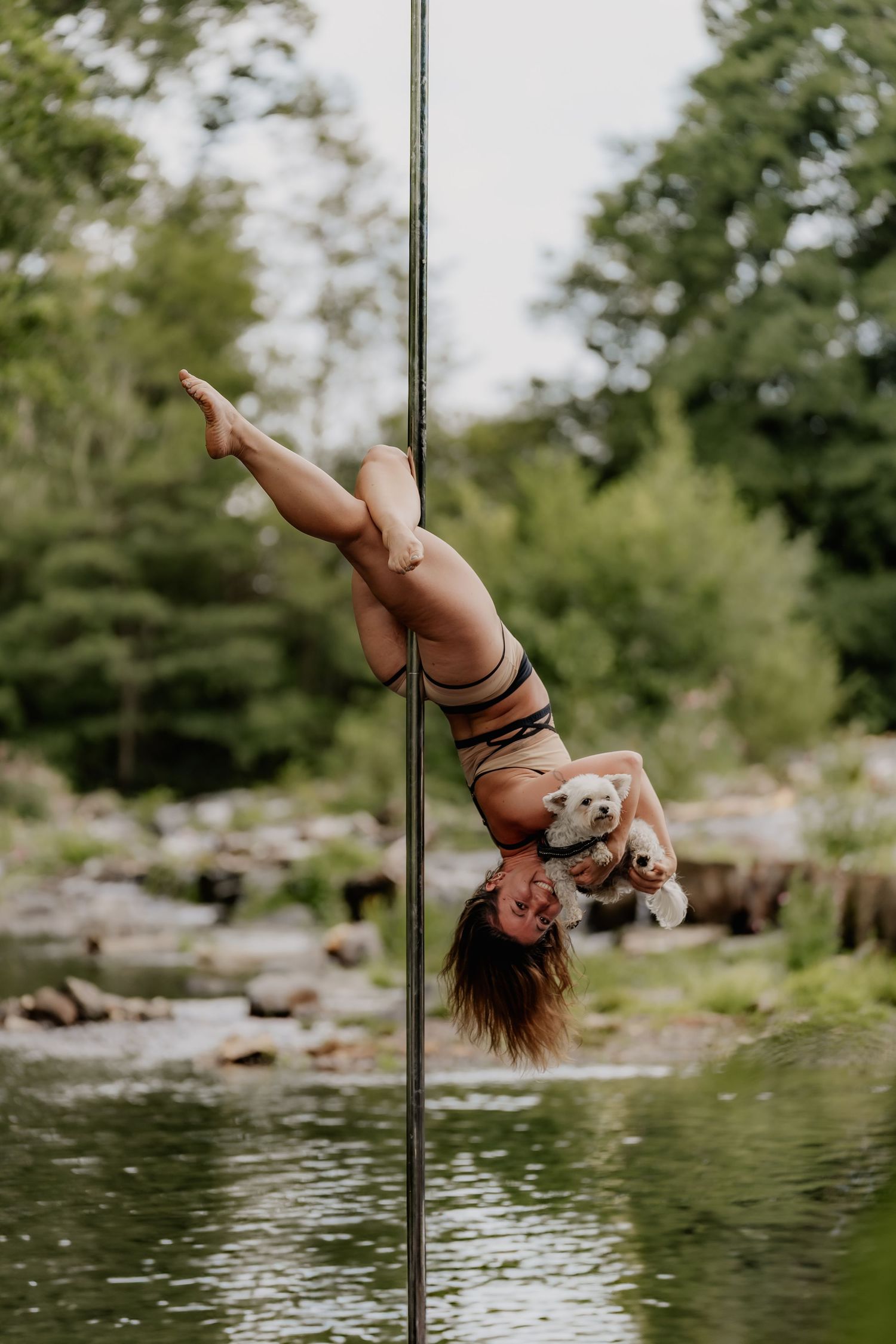 A woman performs an inverted pole dance move outdoors near a rocky river, holding a small white dog while upside down.