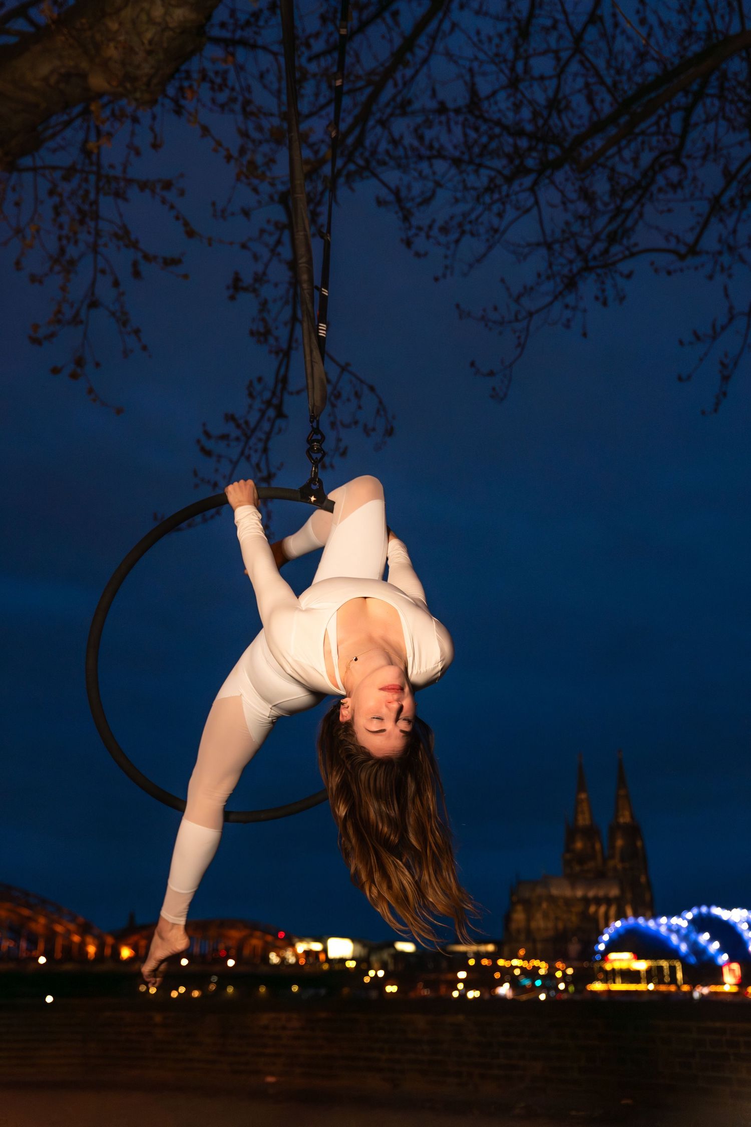 Aerial hoop dancer in white performs upside-down at night with city lights and cathedral silhouette in background.
