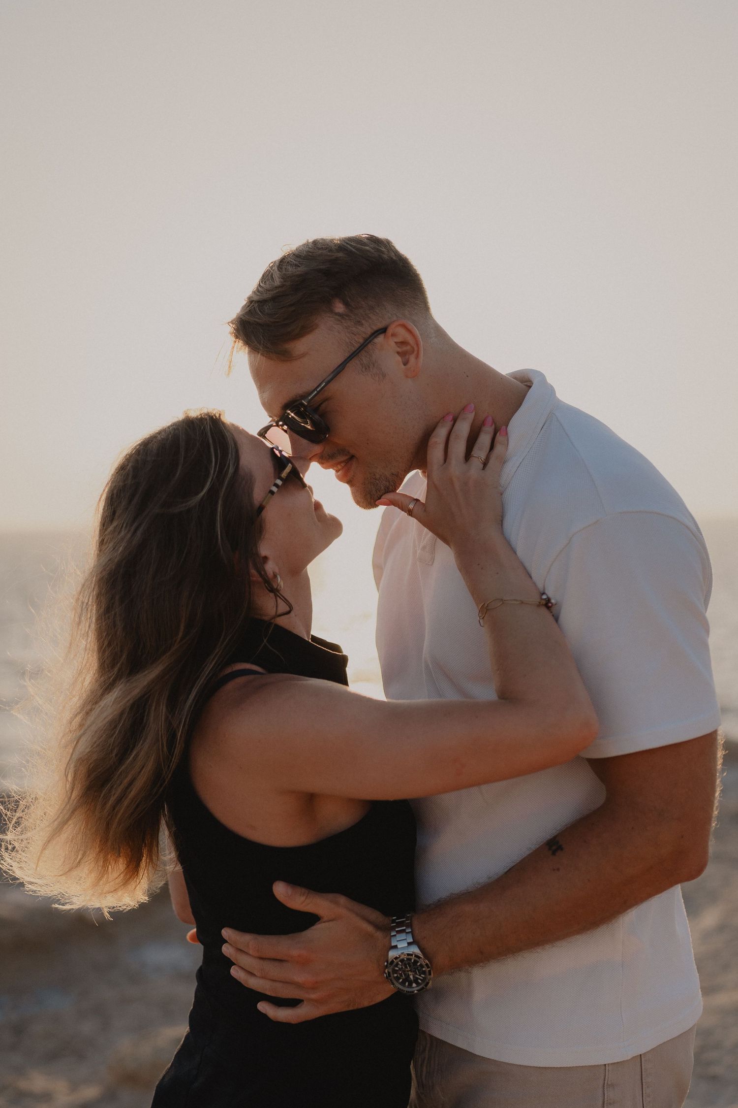 A romantic couple embracing and nearly kissing at sunset on a beach, both wearing sunglasses.