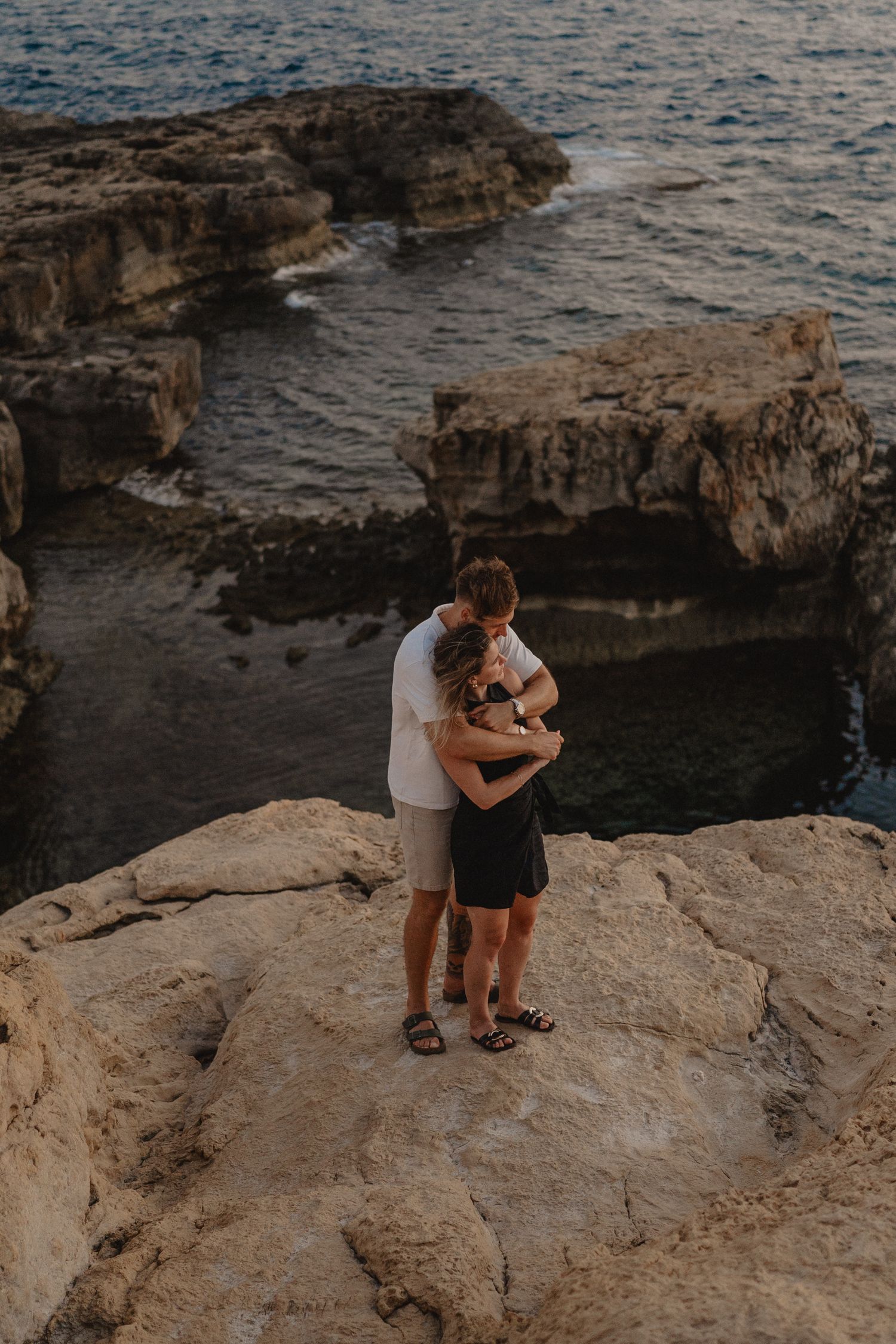 Couple embracing on rocky coastal cliffs overlooking the ocean at sunset.