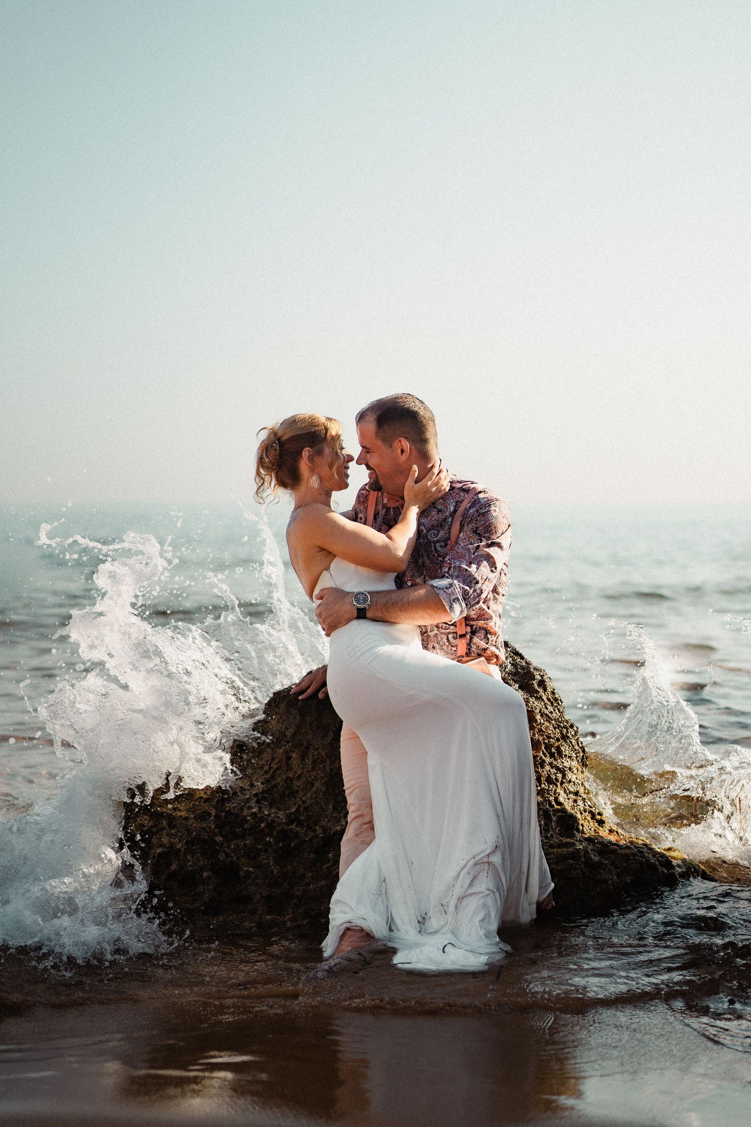 Couple embracing on a rock in the ocean waves, woman in white dress and man in floral shirt, romantic beach photo.