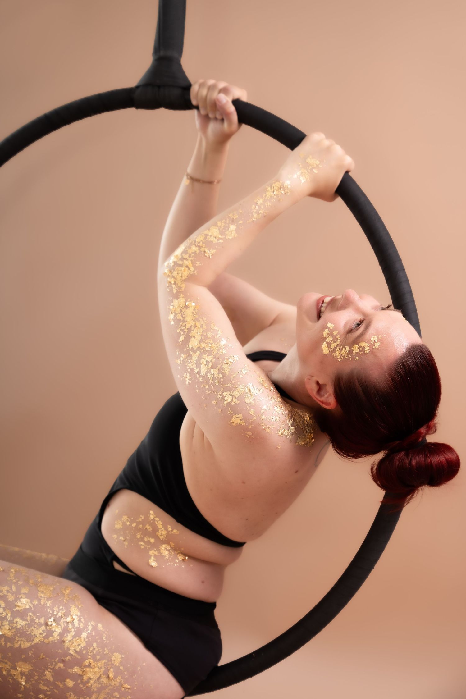 Woman with gold leaf body art performs aerial hoop routine in black leotard against beige background.