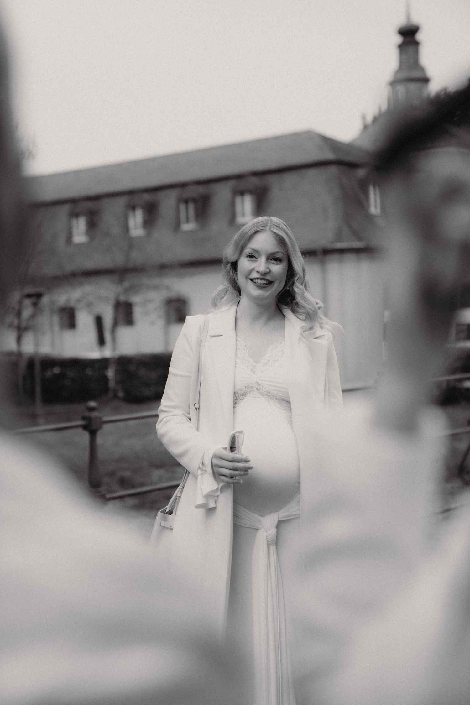 Black and white photo of smiling pregnant woman in white dress and coat outdoors.