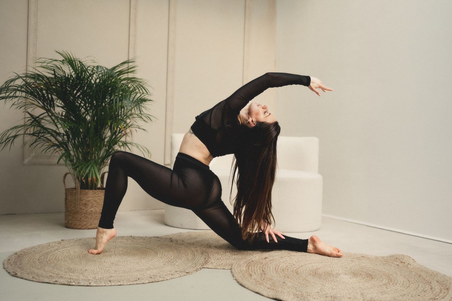 Woman in black activewear performing a yoga lunge pose with arm raised in a bright studio with plants.