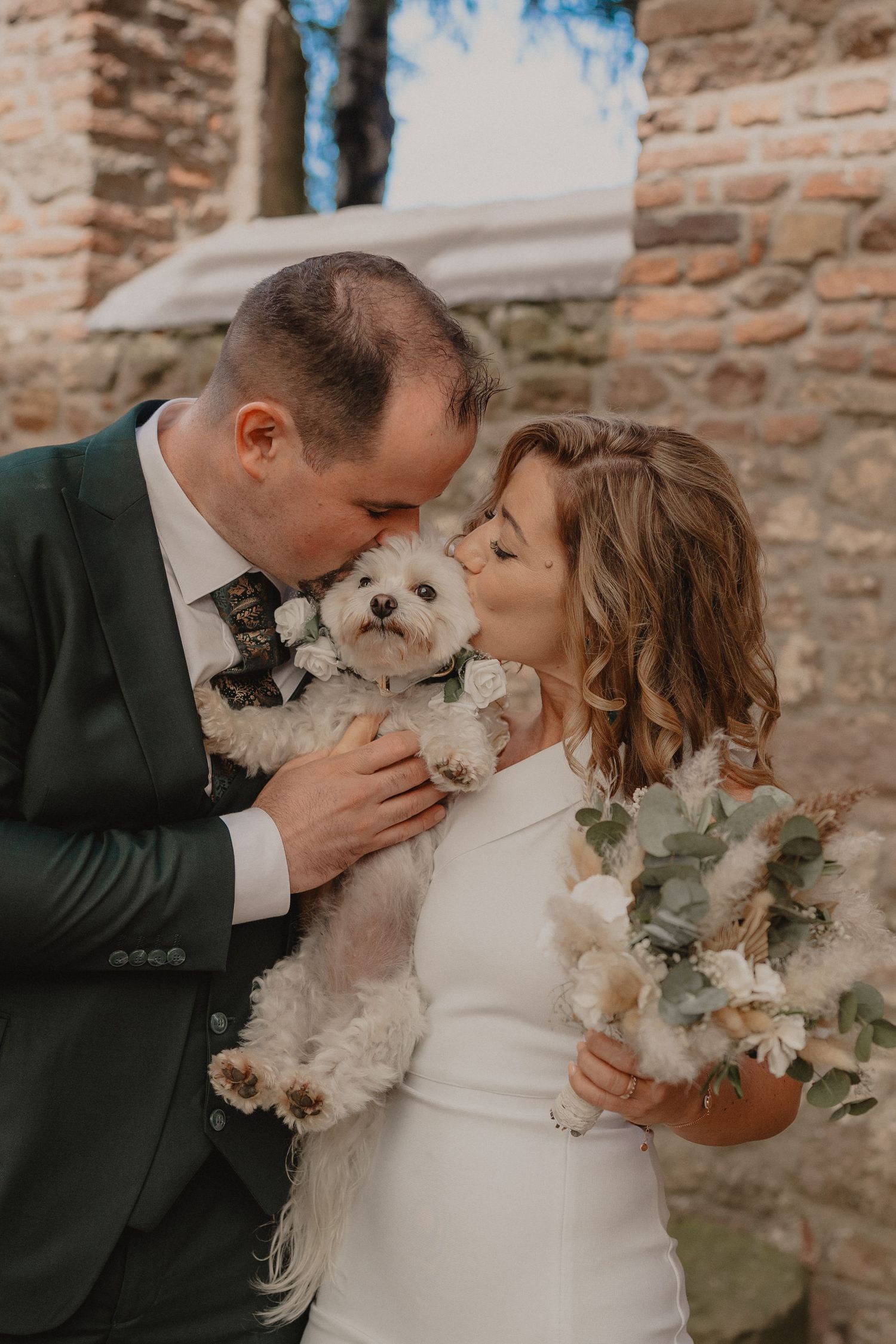 Newlyweds sharing romantic moment with white floral bouquet against rustic brick wall backdrop.