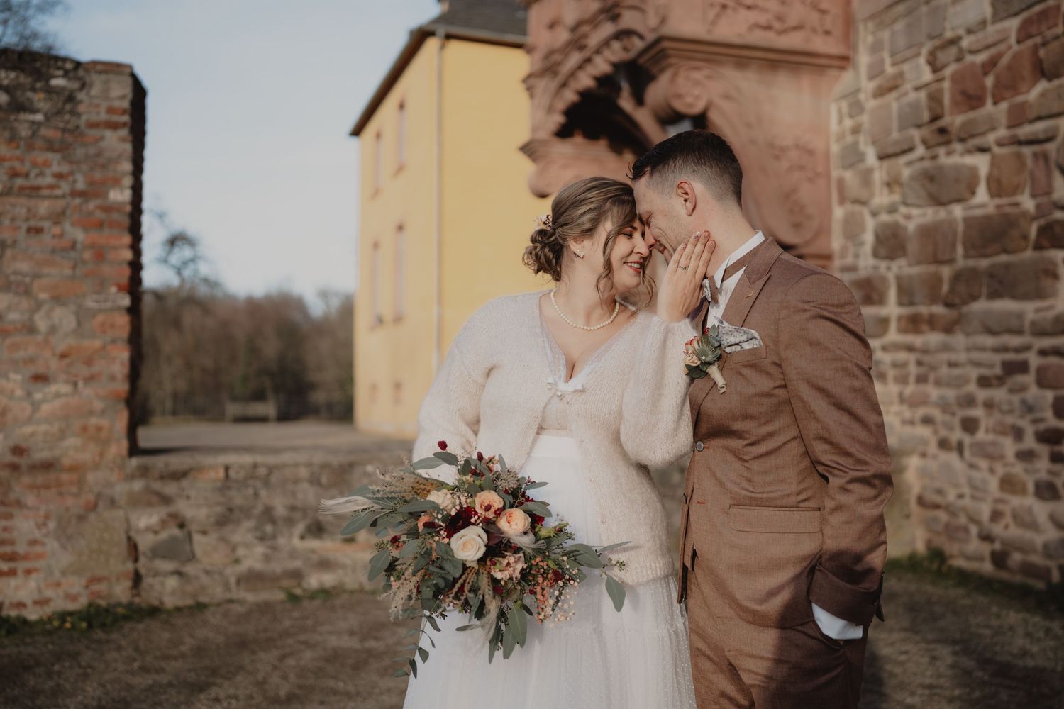 Romantic outdoor moment against historic stone wall with autumn-toned floral bouquet.