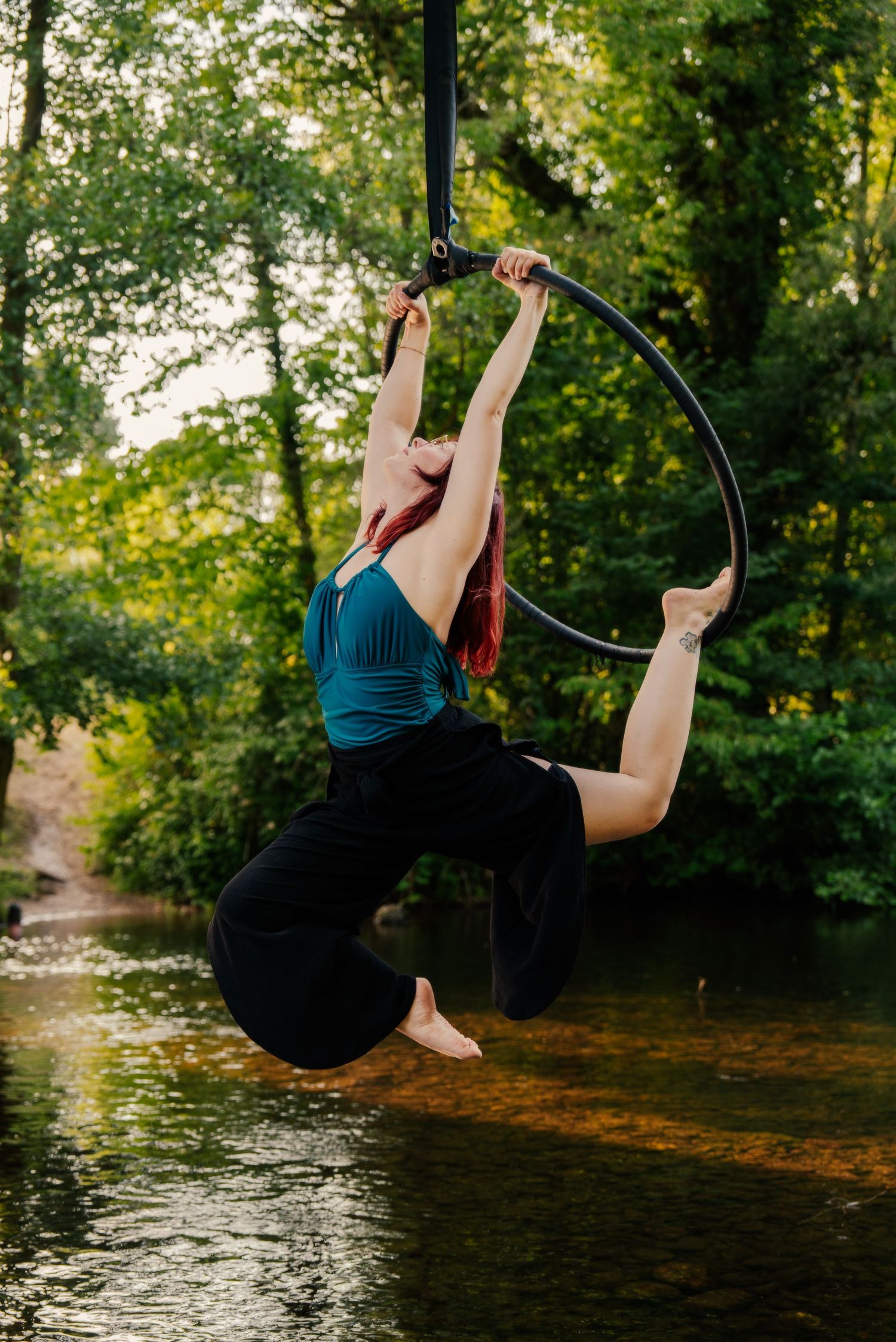 Aerial hoop artist performs graceful pose suspended over a serene river surrounded by lush green trees outdoors.
