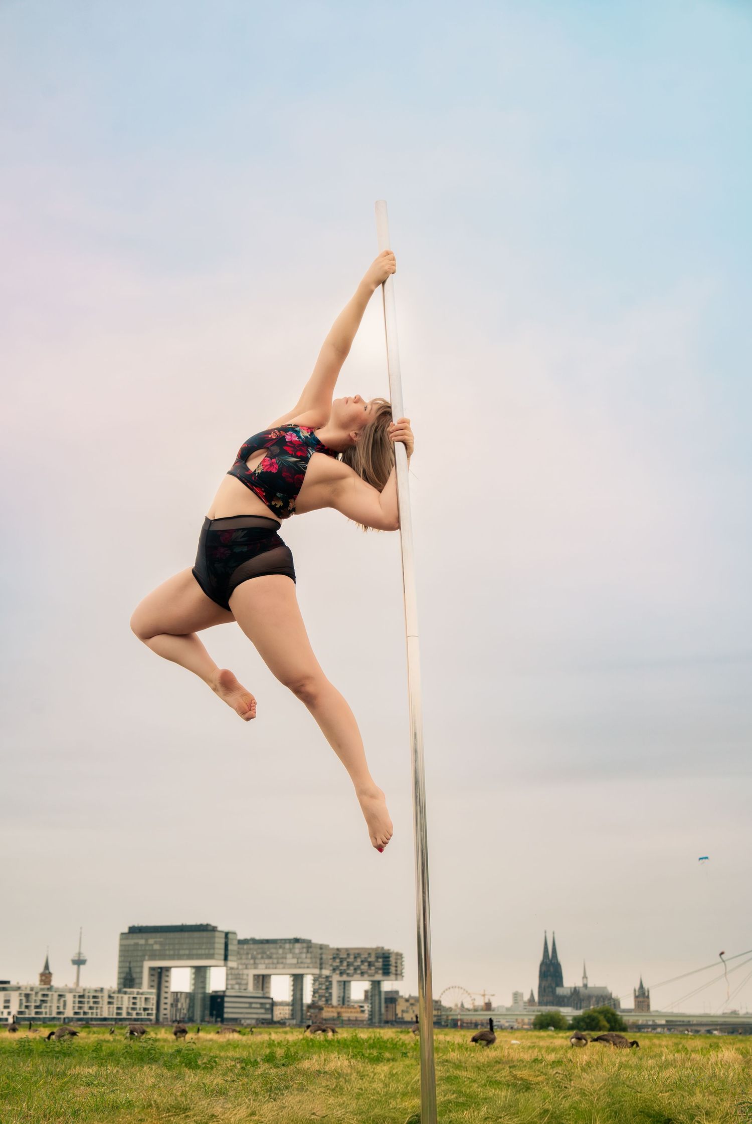 Woman performing pole dance outdoors near Cologne skyline, wearing black shorts and floral top.