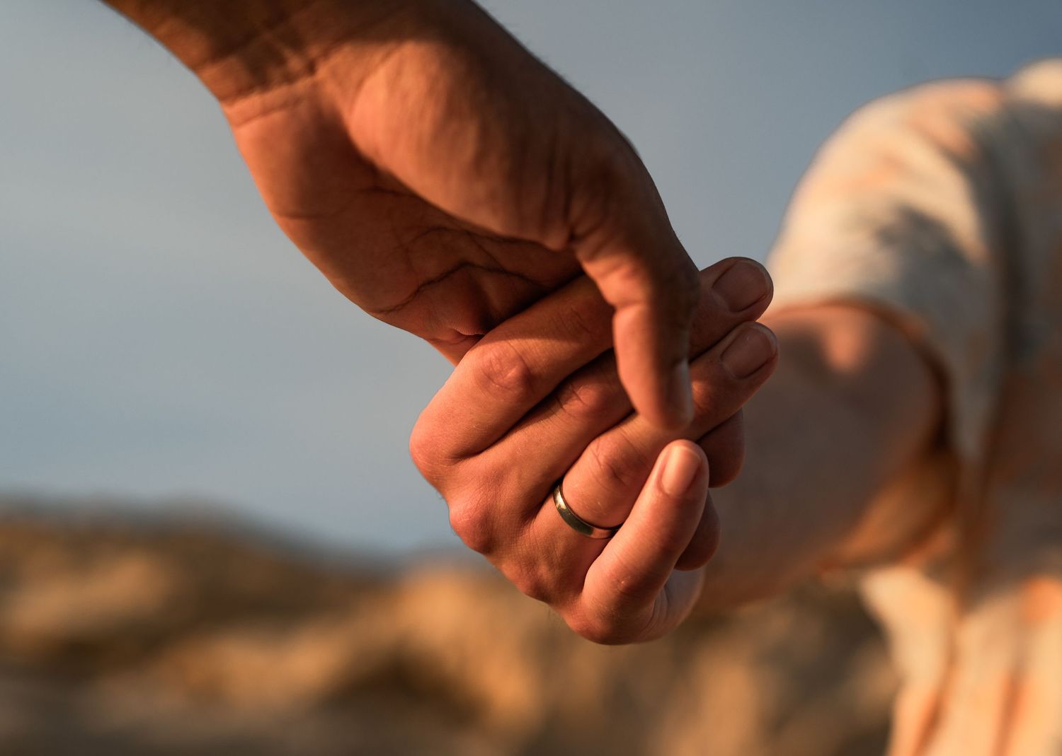 Close-up of two people holding hands, one wearing a black ring, against a warm sunset outdoor background.