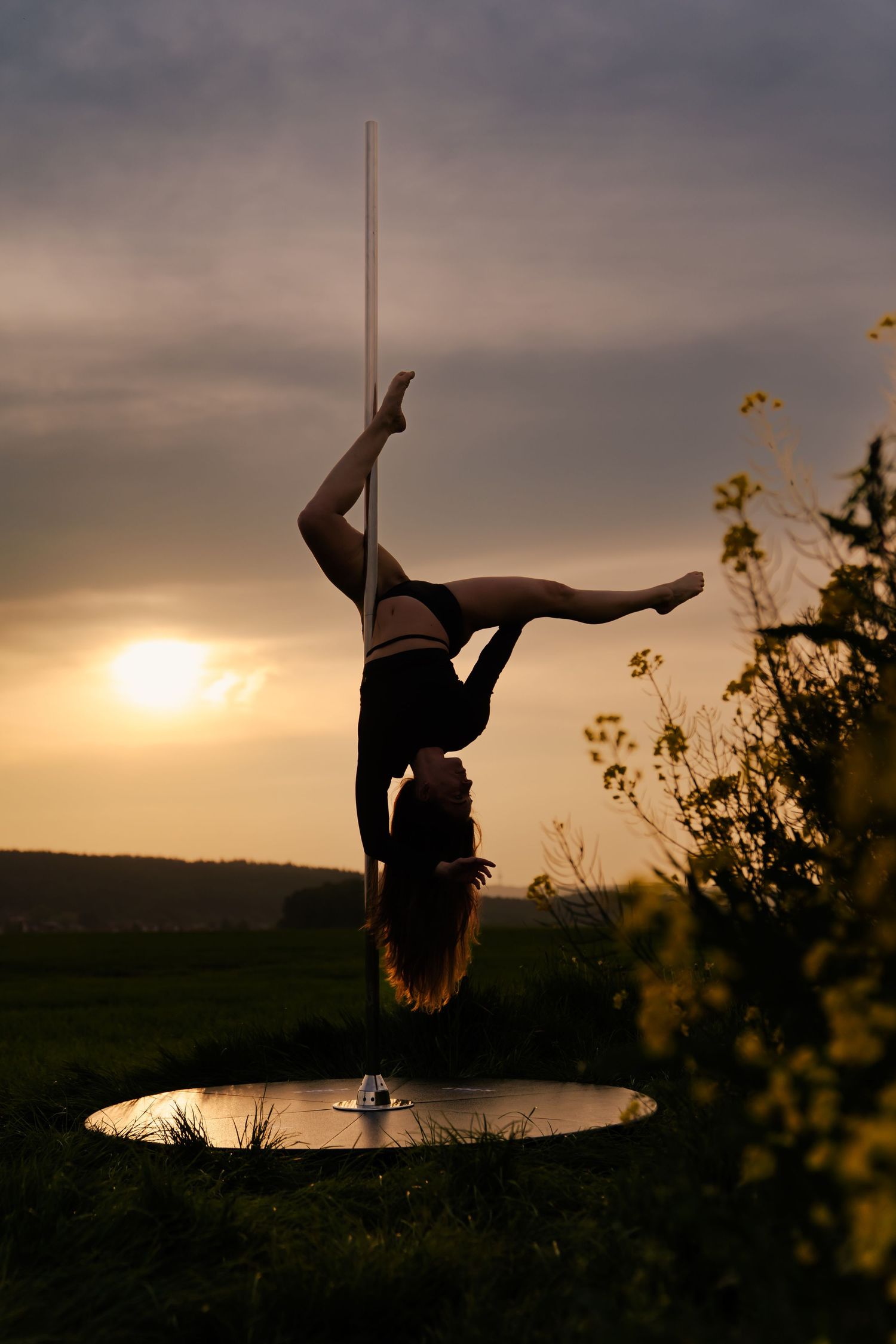 Silhouette of a pole dancer performing a handstand at sunset in an outdoor field setting.