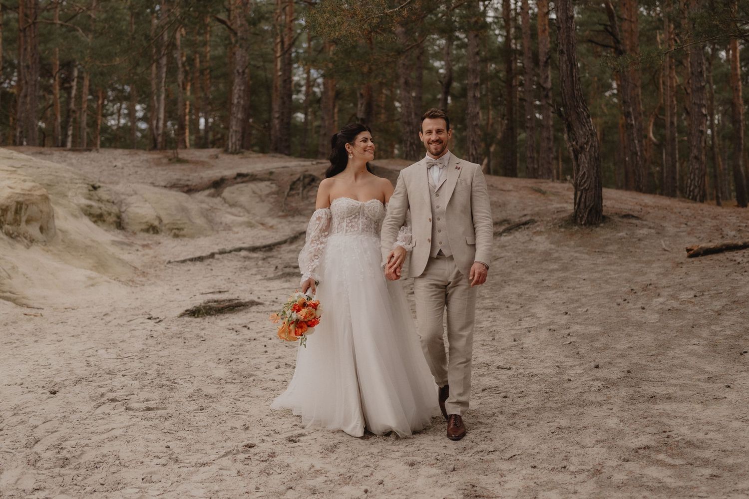 Wedding couple walks hand in hand through a sandy forest landscape.