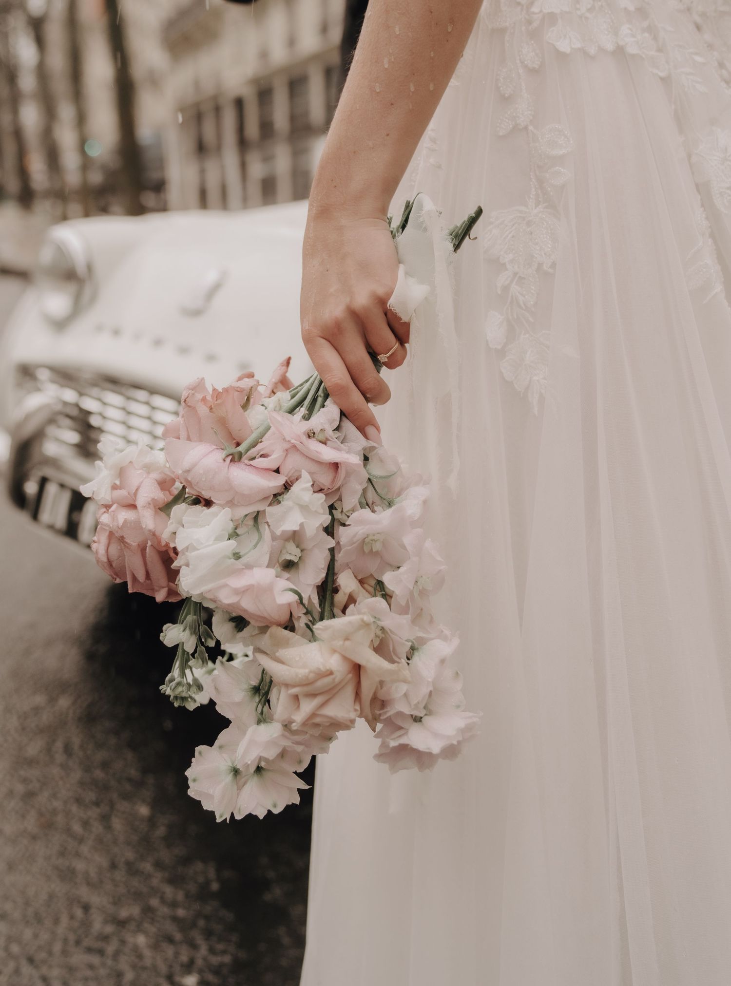 A bride in a white dress holds a delicate bouquet of pink and white flowers against a blurred background.