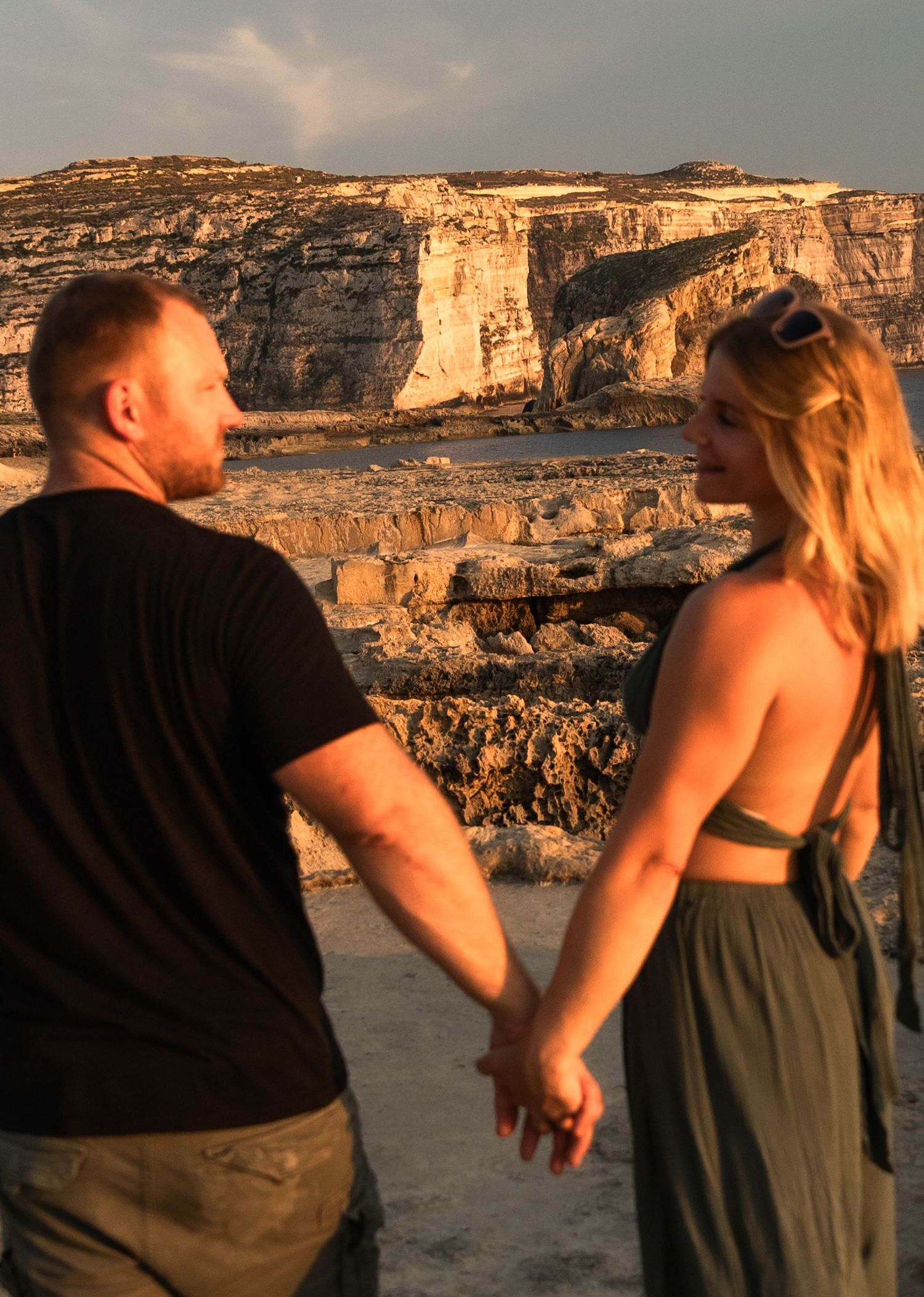 Couple holding hands while admiring scenic coastal cliffs at sunset, woman in green dress with sunglasses.