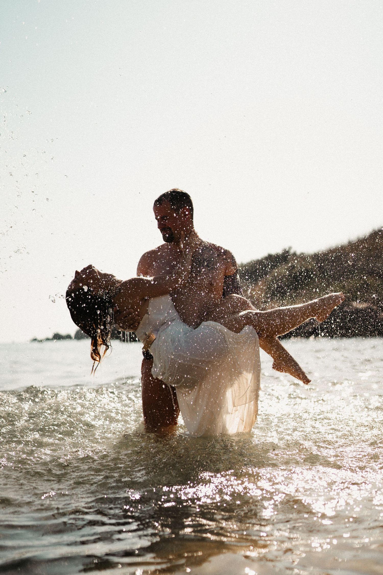 A man carries a woman in a white dress through ocean waves on a sunny beach, water splashing around them.