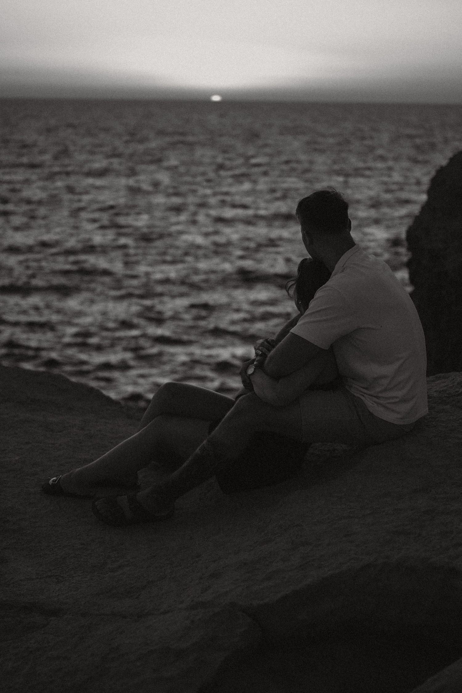 Black and white photo of a couple sitting on rocks, embracing while watching the sunset over the ocean.