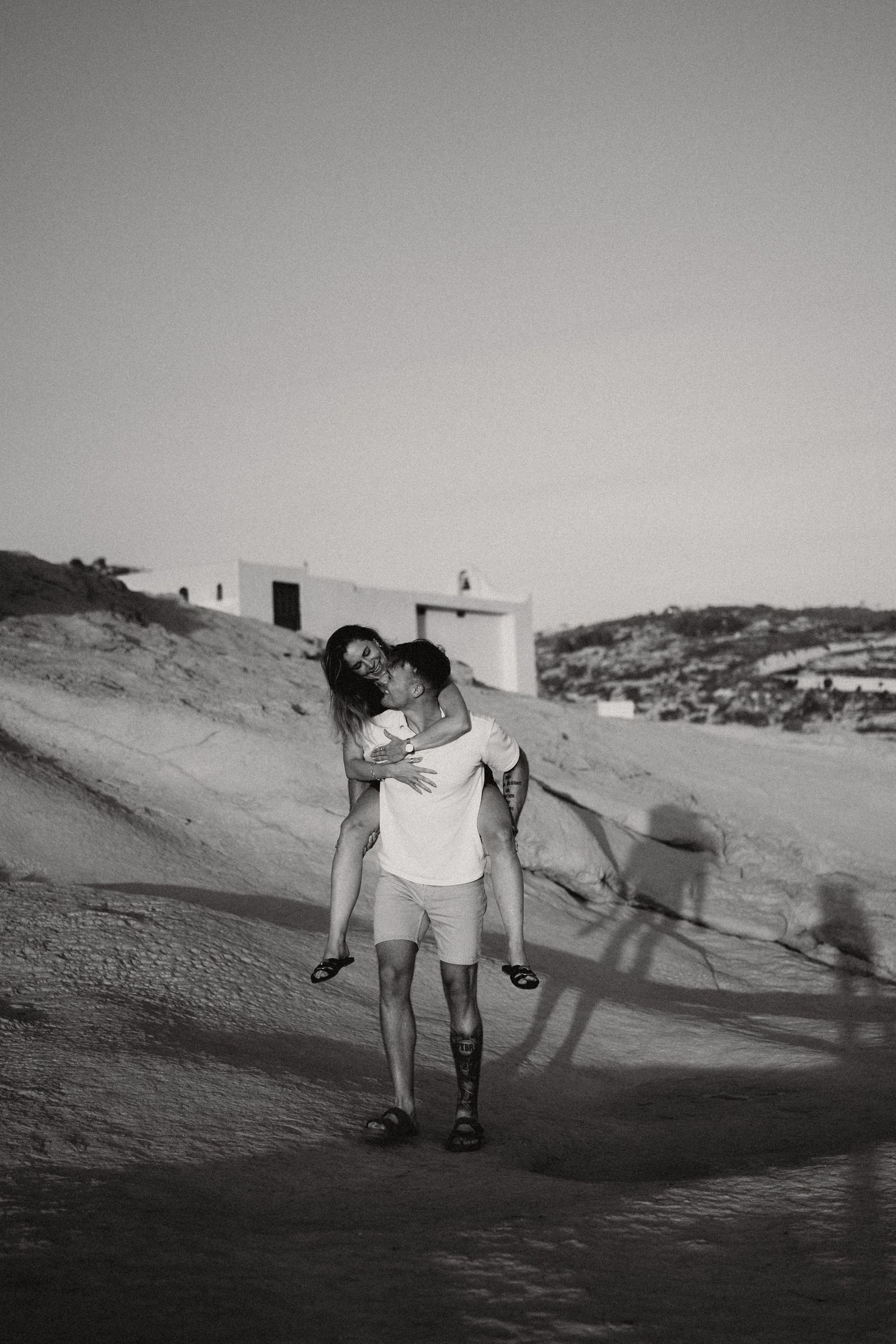 Black and white photo of a couple embracing on a sandy path near white buildings in a Mediterranean coastal setting.