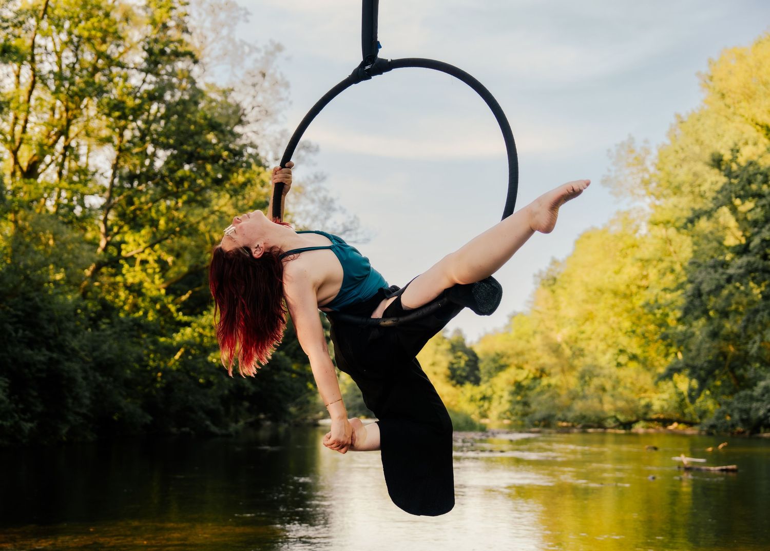 A woman performs aerial hoop acrobatics over a river surrounded by lush green trees outdoors.